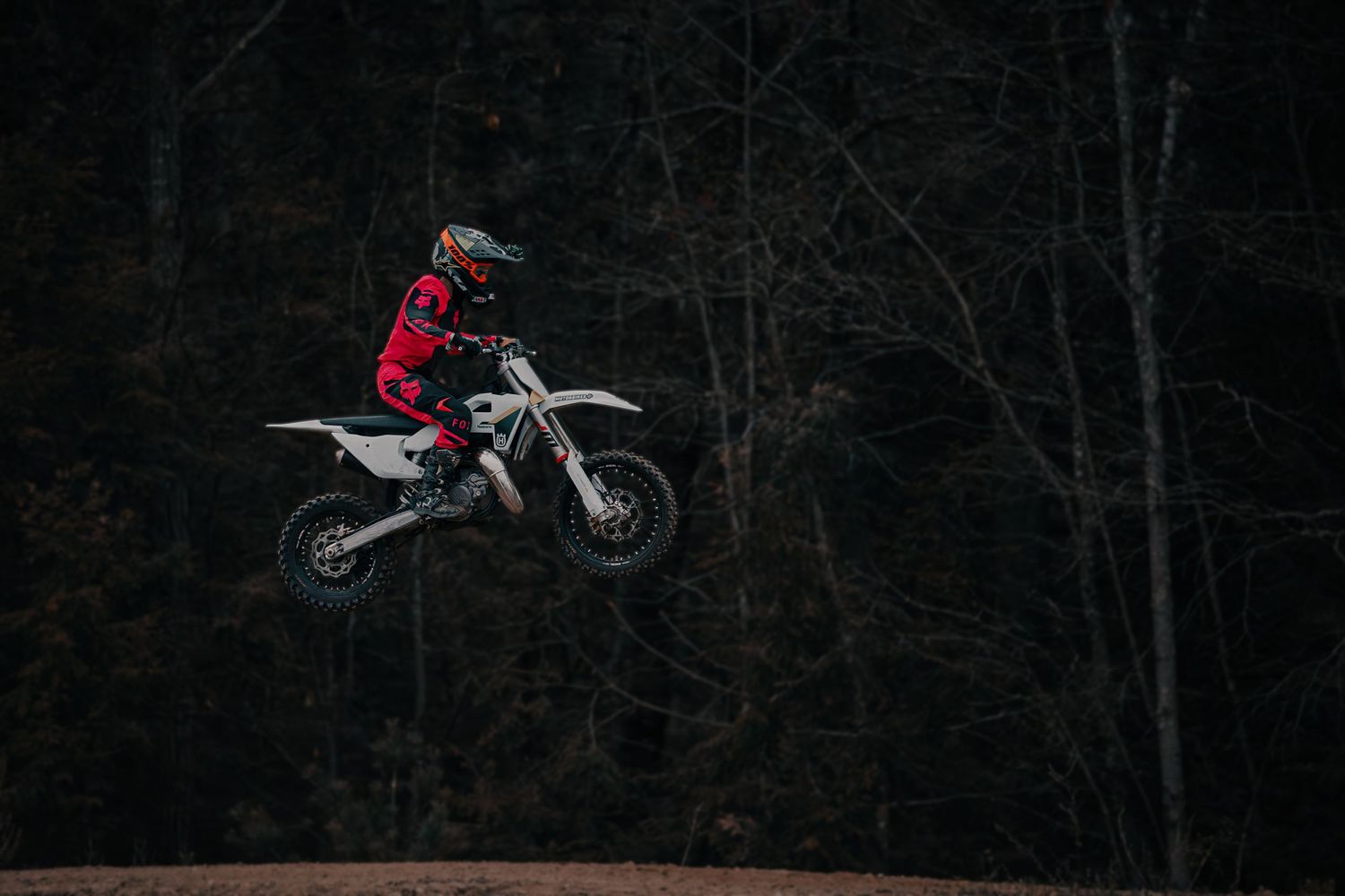 Motocross rider in red gear performs a high jump on a white dirt bike against a dark forest background.