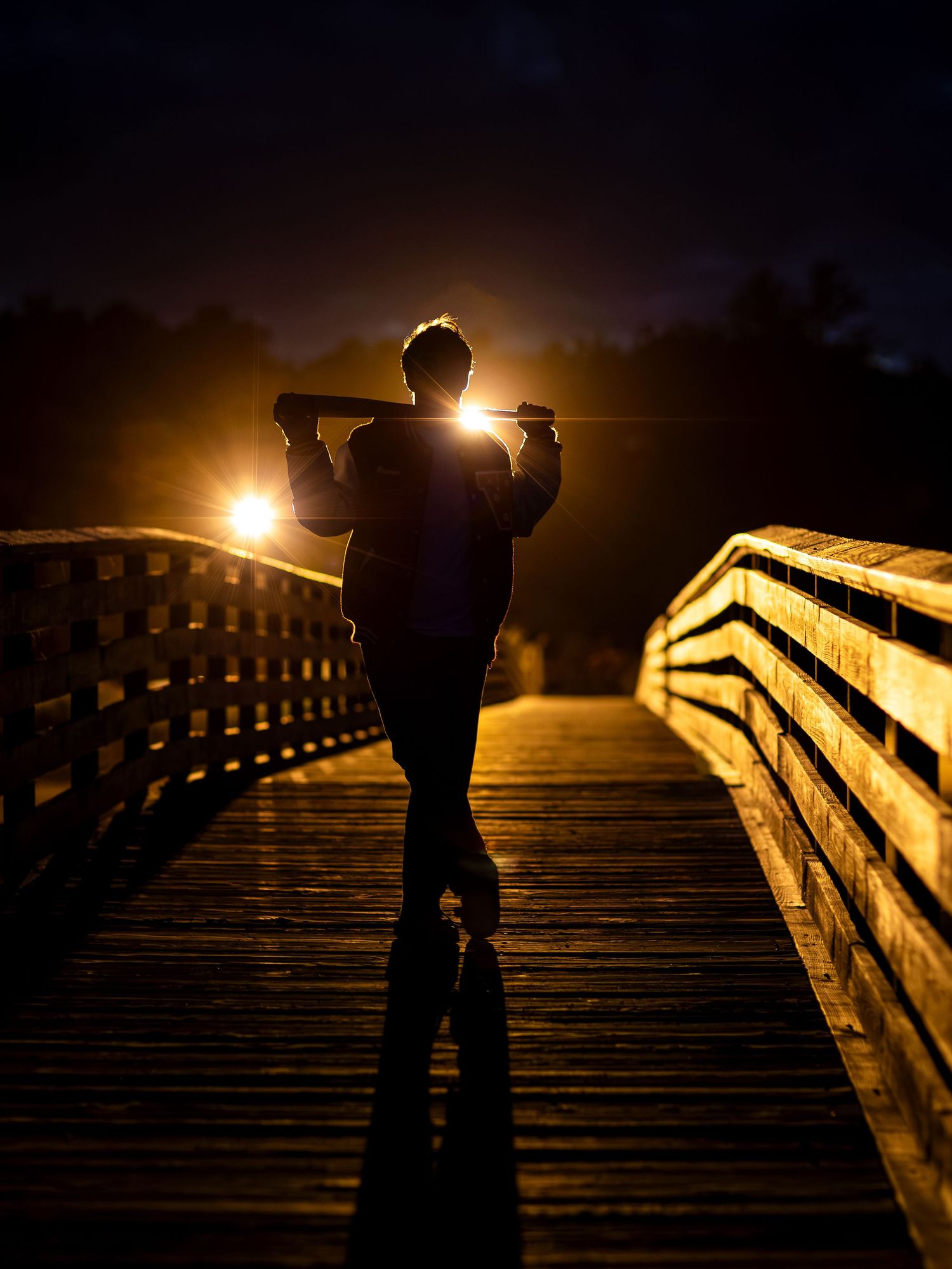 A silhouetted figure stands on a wooden boardwalk at night illuminated by warm glowing lights in the darkness.