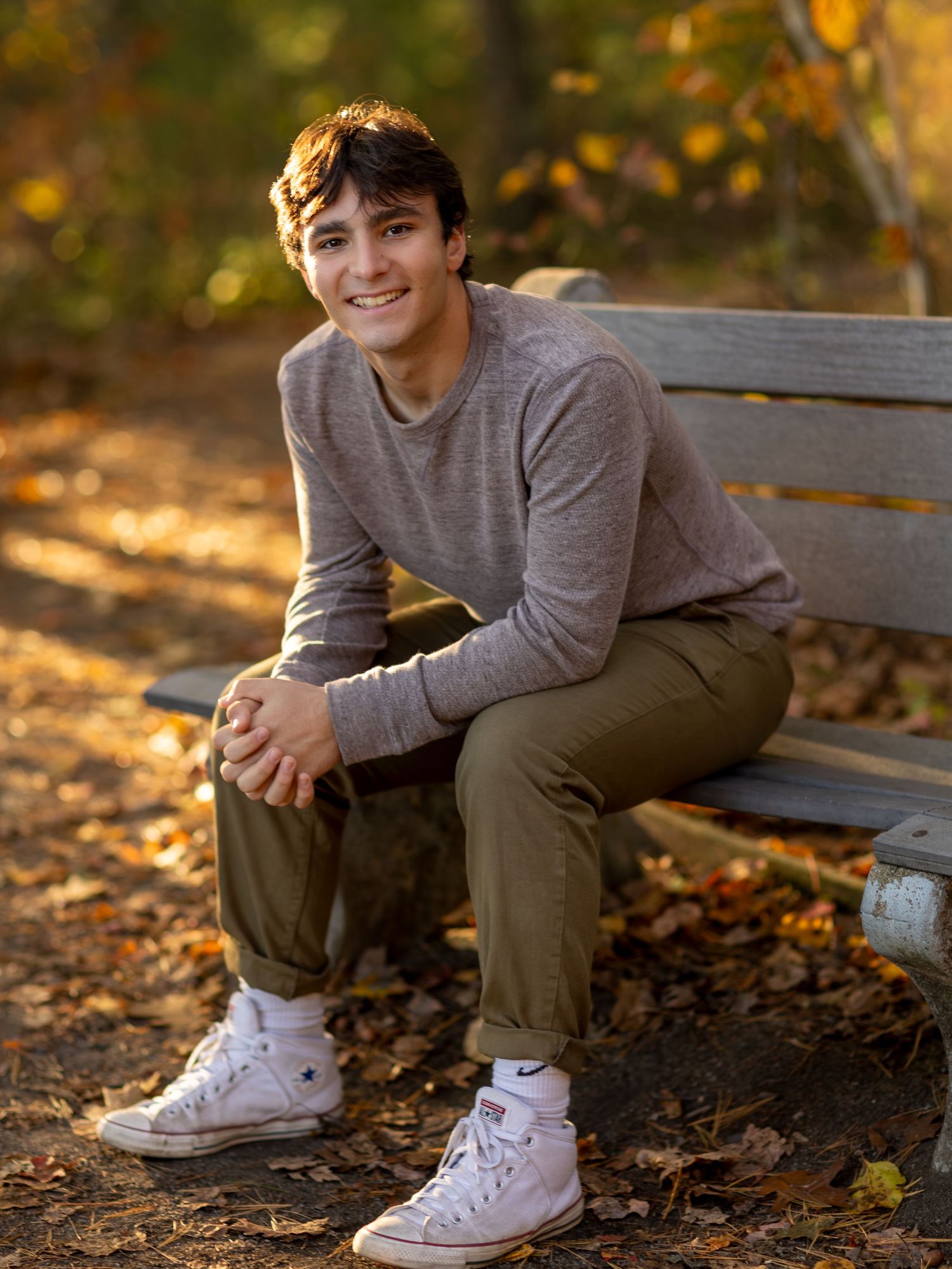 A graduating Senior sits on a wooden bench surrounded by fallen autumn leaves while wearing a gray sweater and white sneakers.