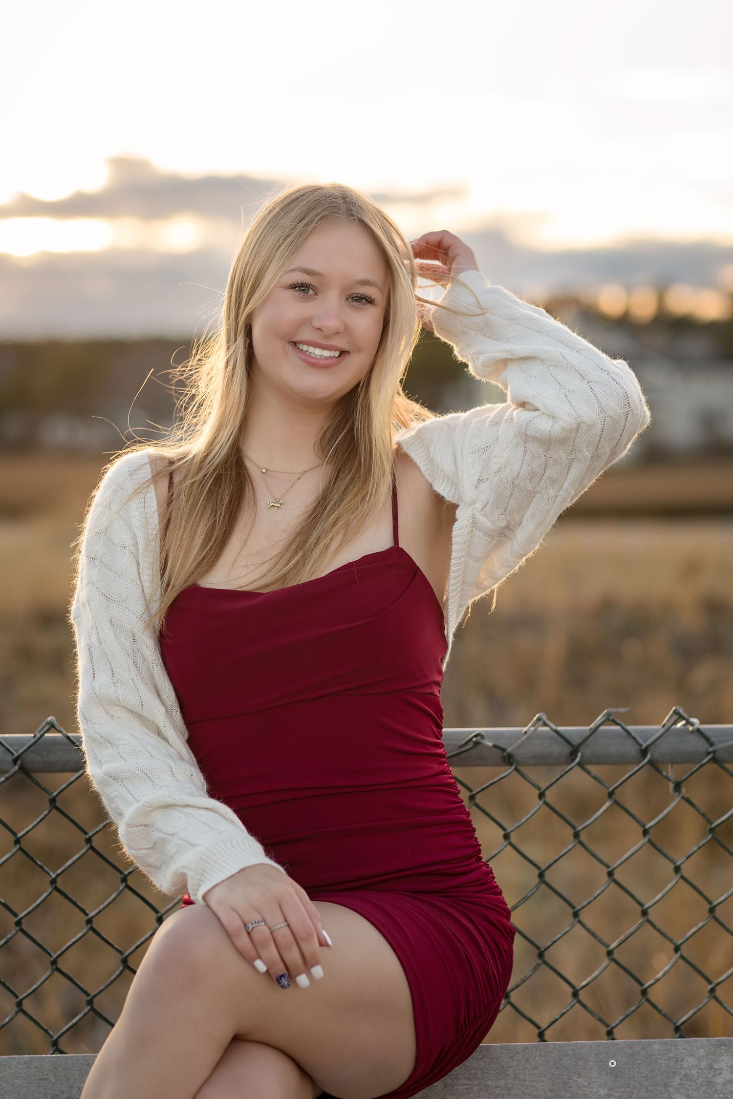 A graduating senior in red dress and white sweater sits on chain link fence at sunset.