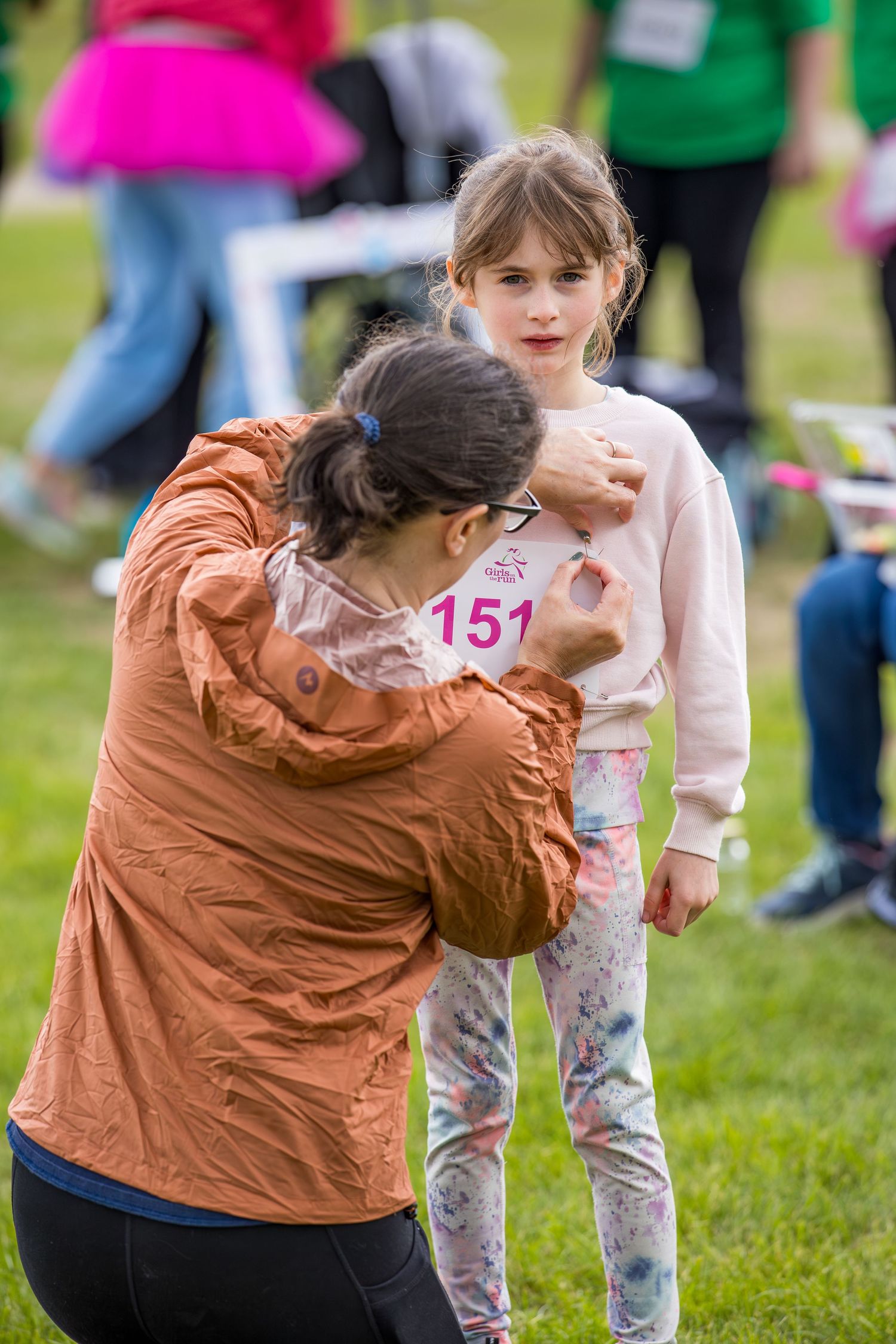Adult pinning race number 151 on a young girl at an outdoor charity fun run event on a green field.
