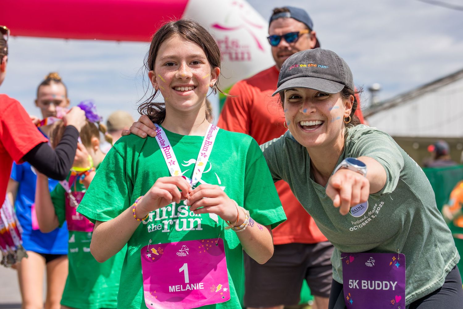 Two girls in green Girls on the Run shirts smile proudly displaying medals at a 5K race finish line.