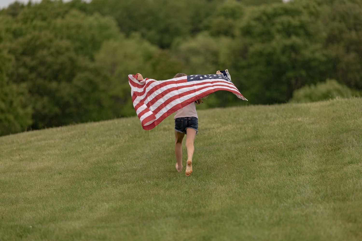 Child running up a green hill holding a large American flag billowing in the wind with trees in background.