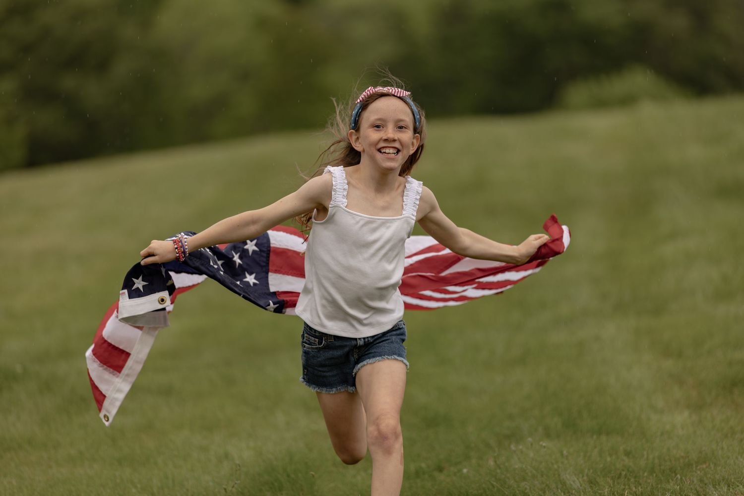 A joyful young girl runs through a green field holding an American flag, celebrating patriotic freedom outdoors.
