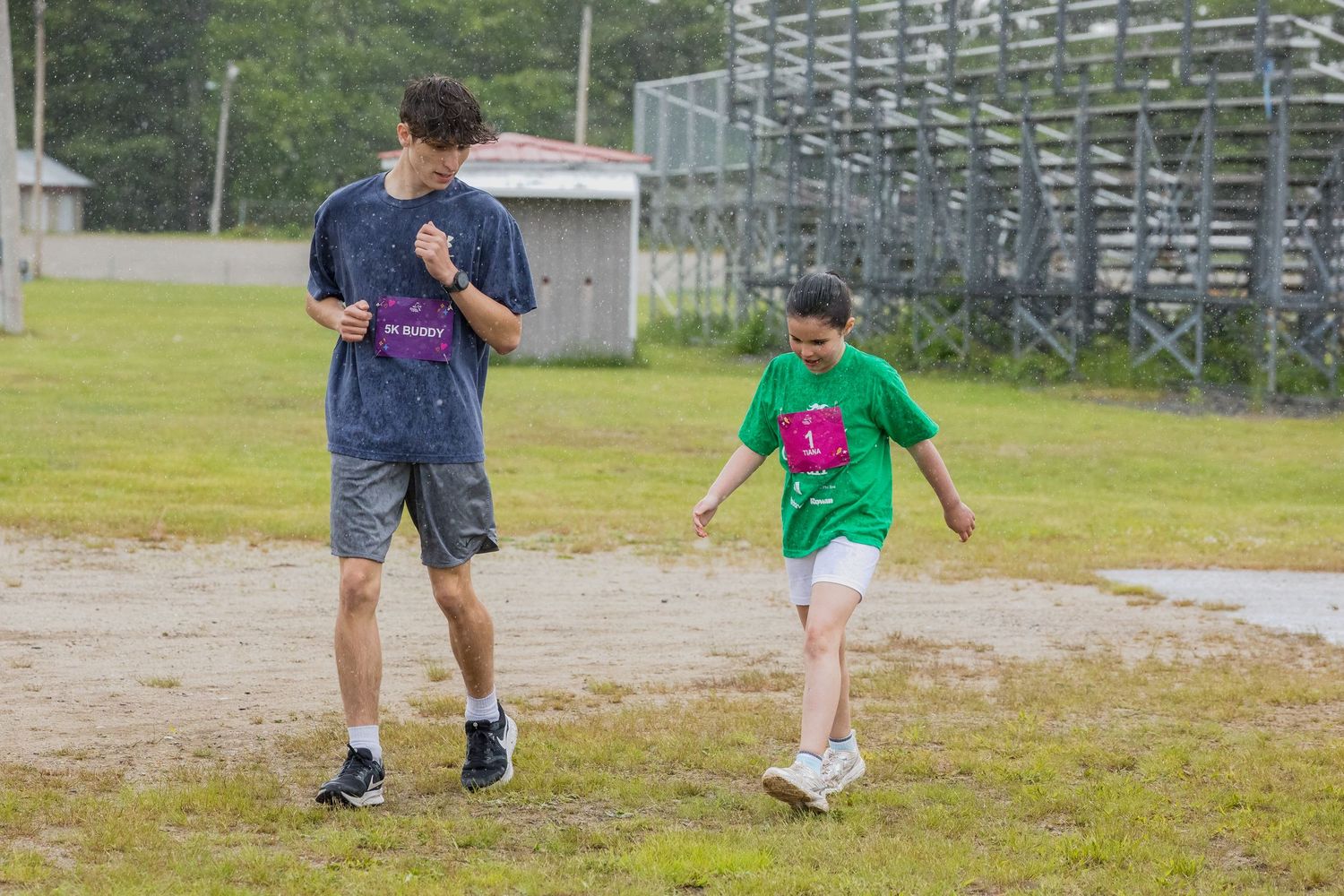 Two kids walking outdoors near bleachers on a rainy day, wearing colorful t-shirts at a summer camp.