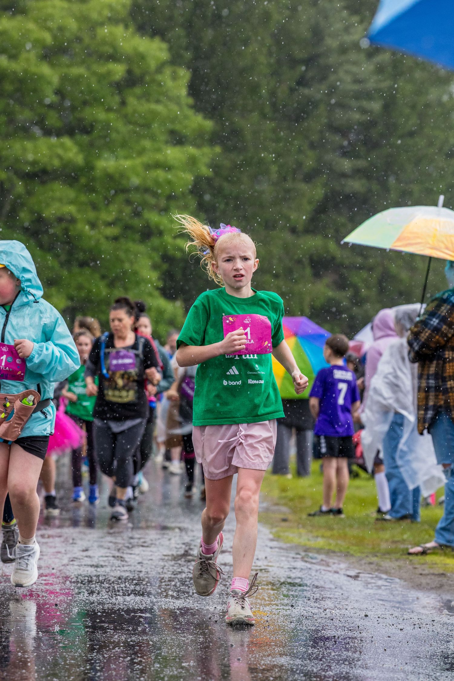 Young girl in green shirt runs through rain puddles during a charity race event with umbrellas in background.