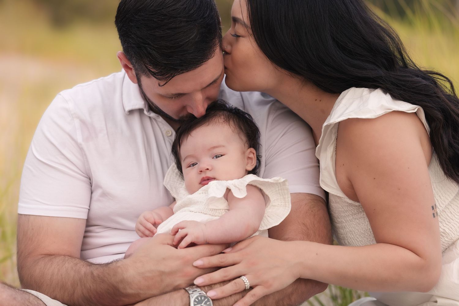 Parents kissing their newborn baby outdoors during a family portrait session in a natural setting.