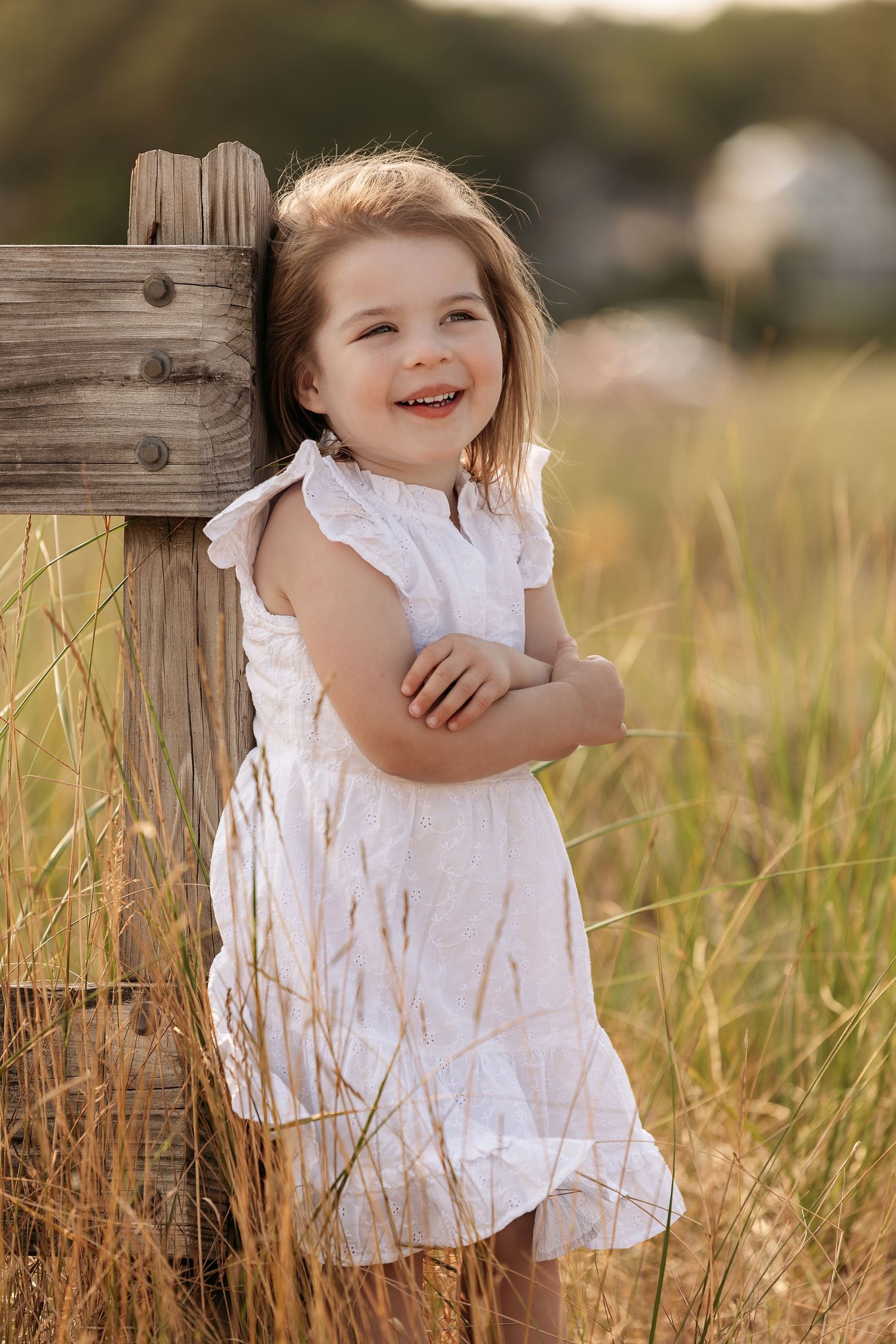 A white sundress-clad figure stands laughing by a wooden fence post in a sunlit field of tall grass.