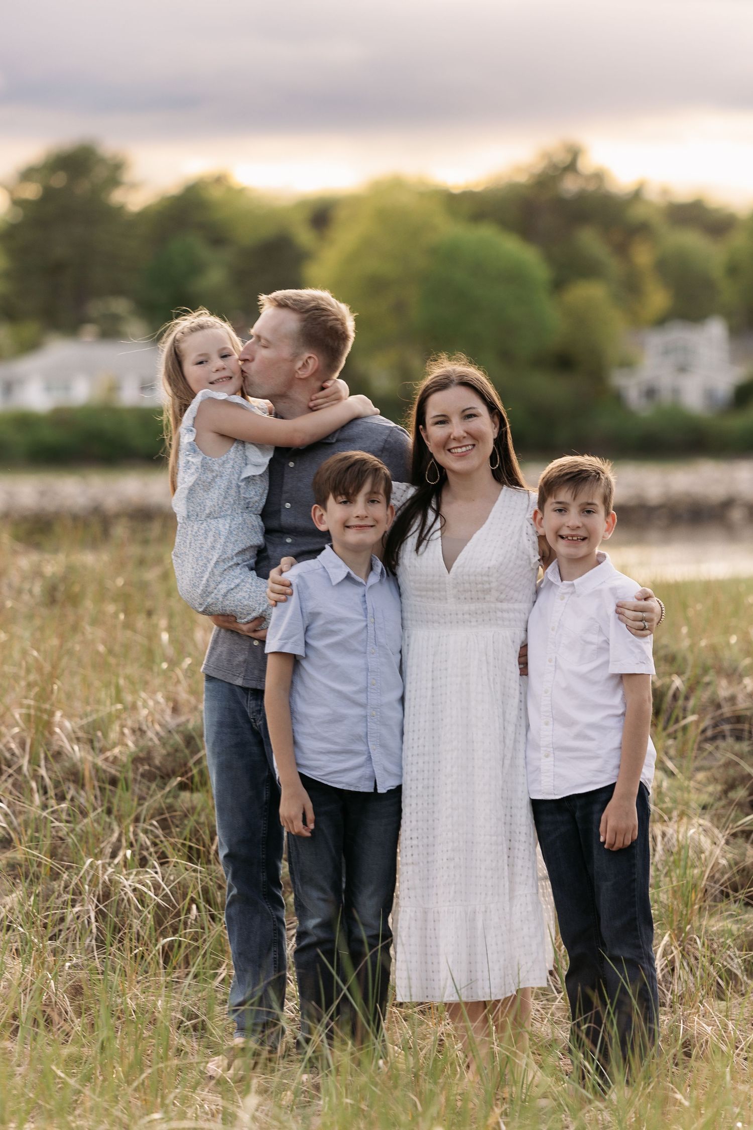 Family poses together in a grassy field at sunset wearing coordinated blue and white outfits.