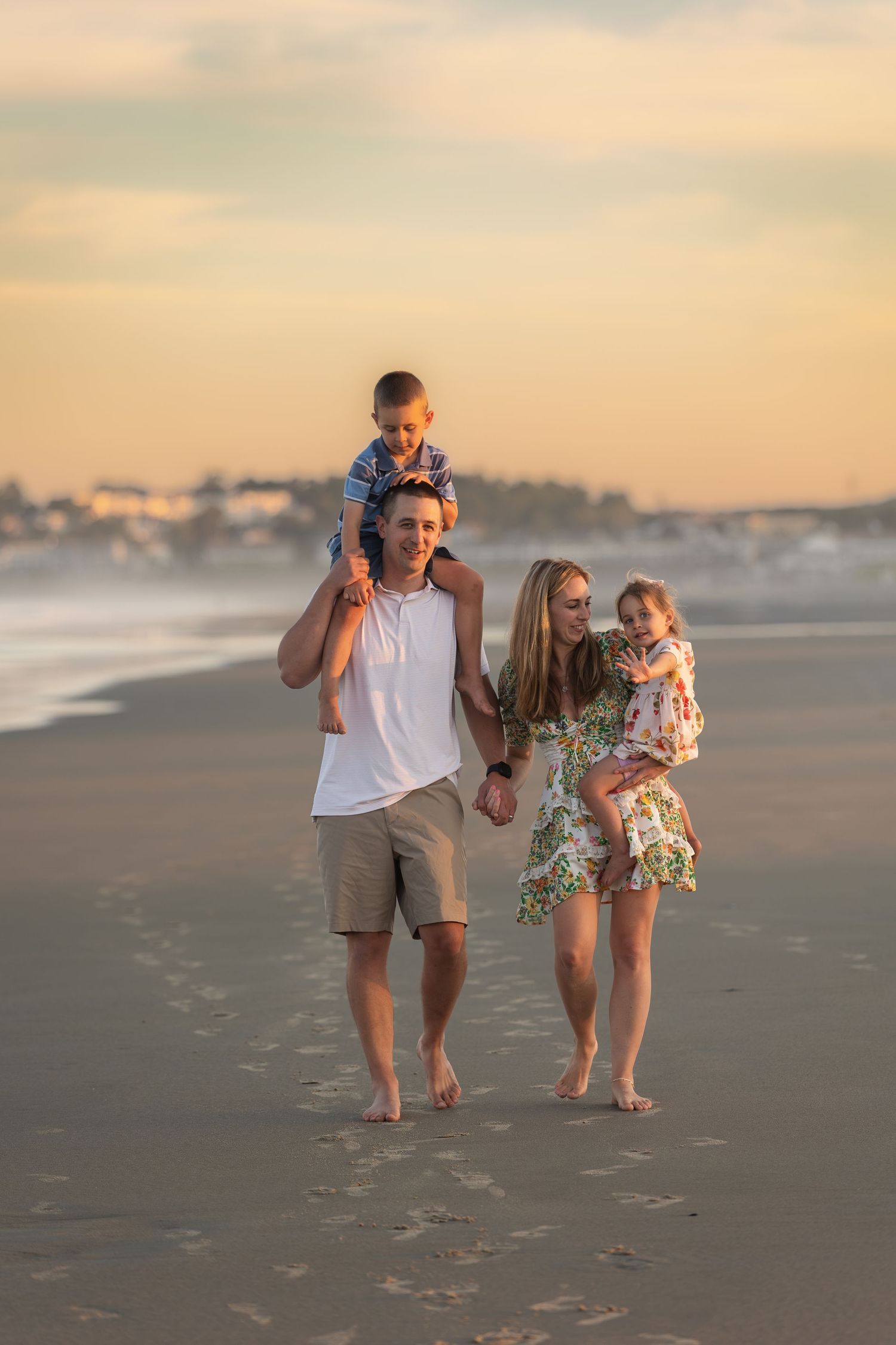 Family of four walking barefoot on a sandy beach at sunset, father carrying son on shoulders, mother holding daughter.