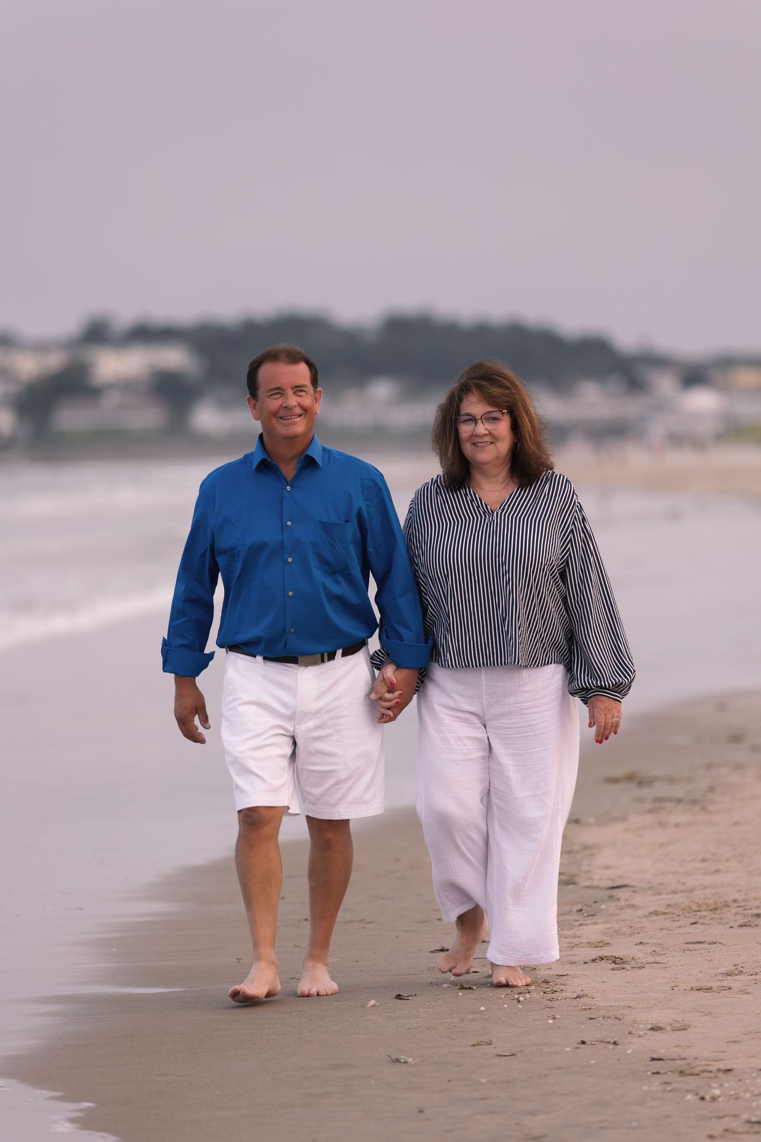 Couple holding hands and walking barefoot along a sandy beach near the ocean.