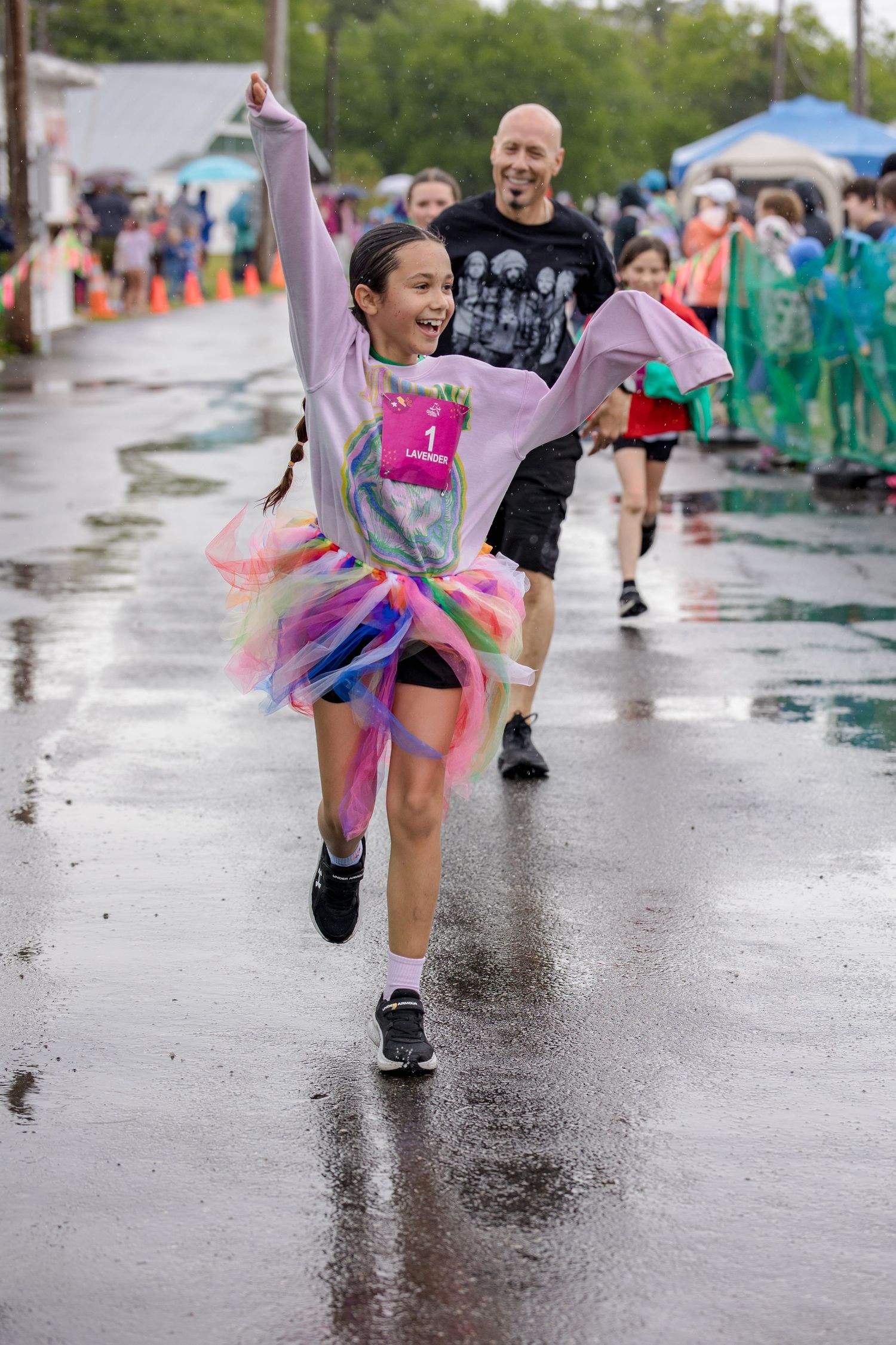 Excited girl in rainbow tutu celebrates finishing 5K race on wet pavement with arms raised.