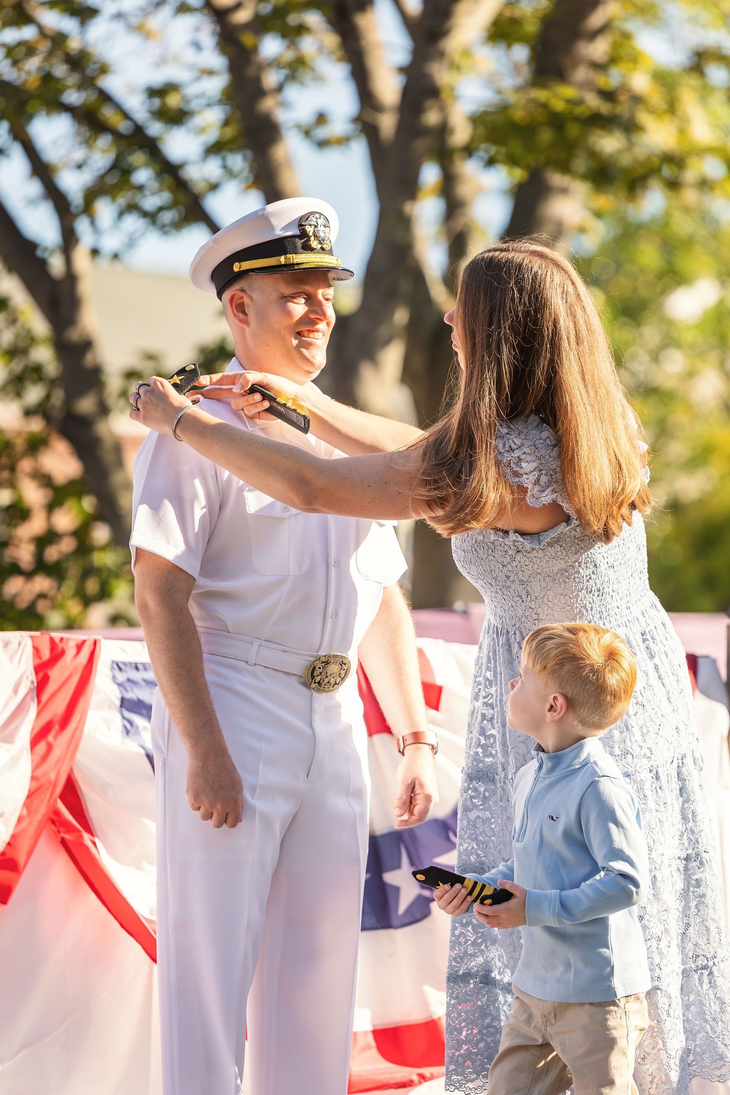 Navy officer in white uniform receives rank insignia from woman outdoors, with young boy nearby and American flags in background.