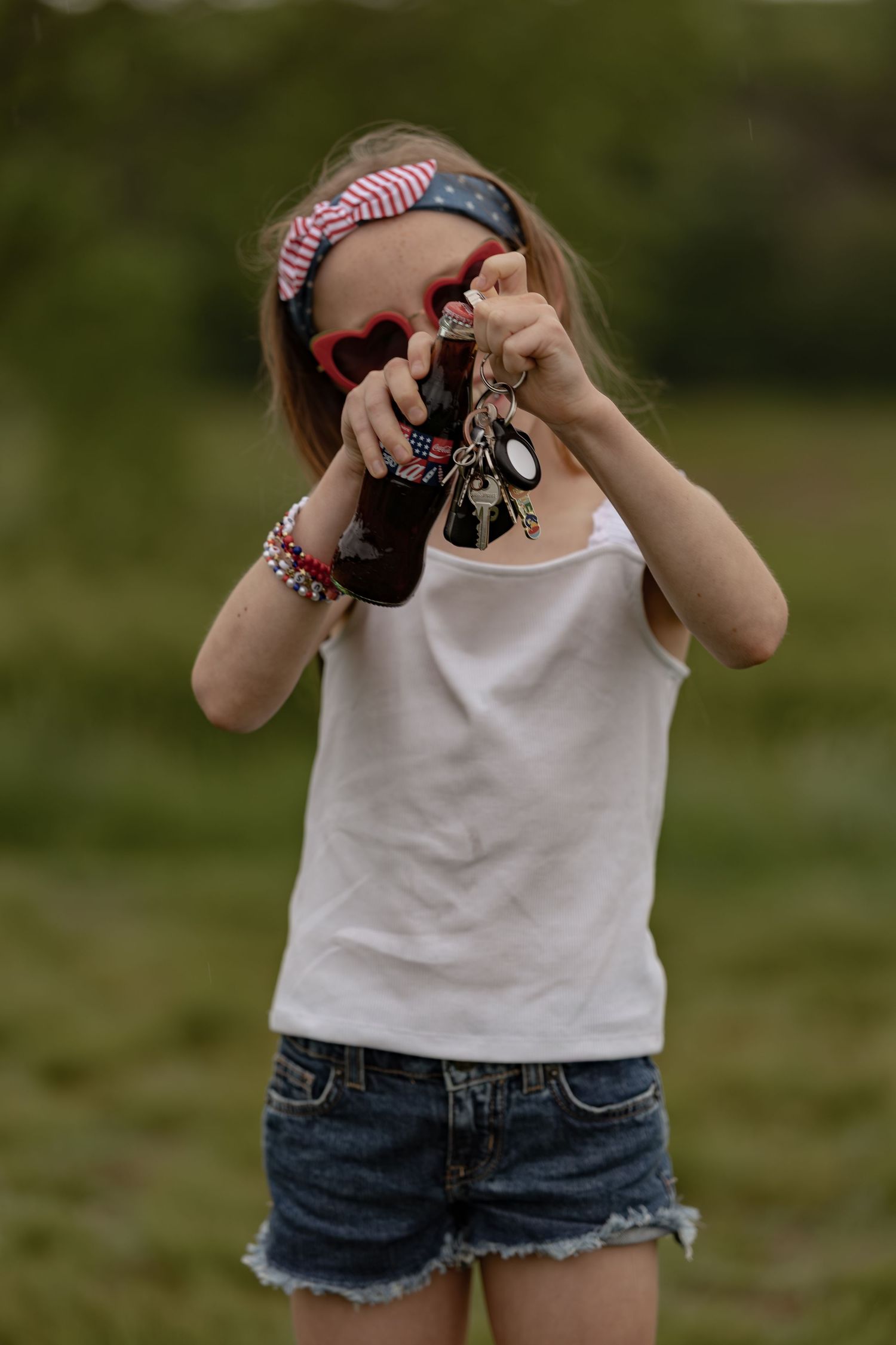 Young girl in white tank top and denim shorts wearing heart sunglasses and red bracelet holding a brown bottle outdoors.