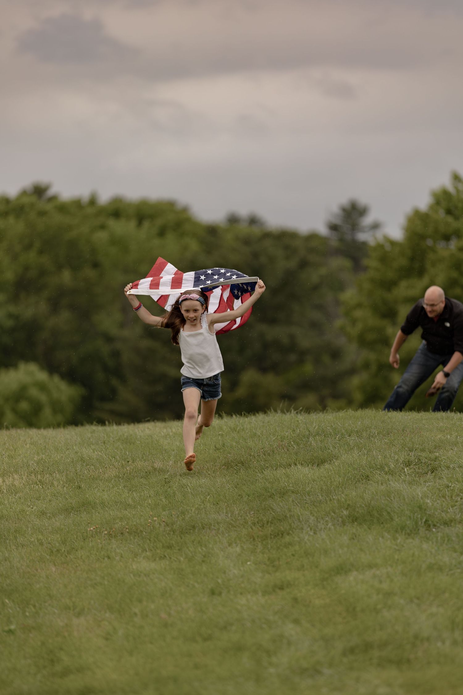 A joyful girl runs across a green hill holding an American flag, with a man and trees visible in the background.
