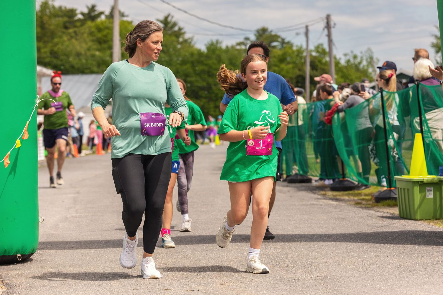 Two women in green shirts running together at an outdoor charity race event on a sunny day.