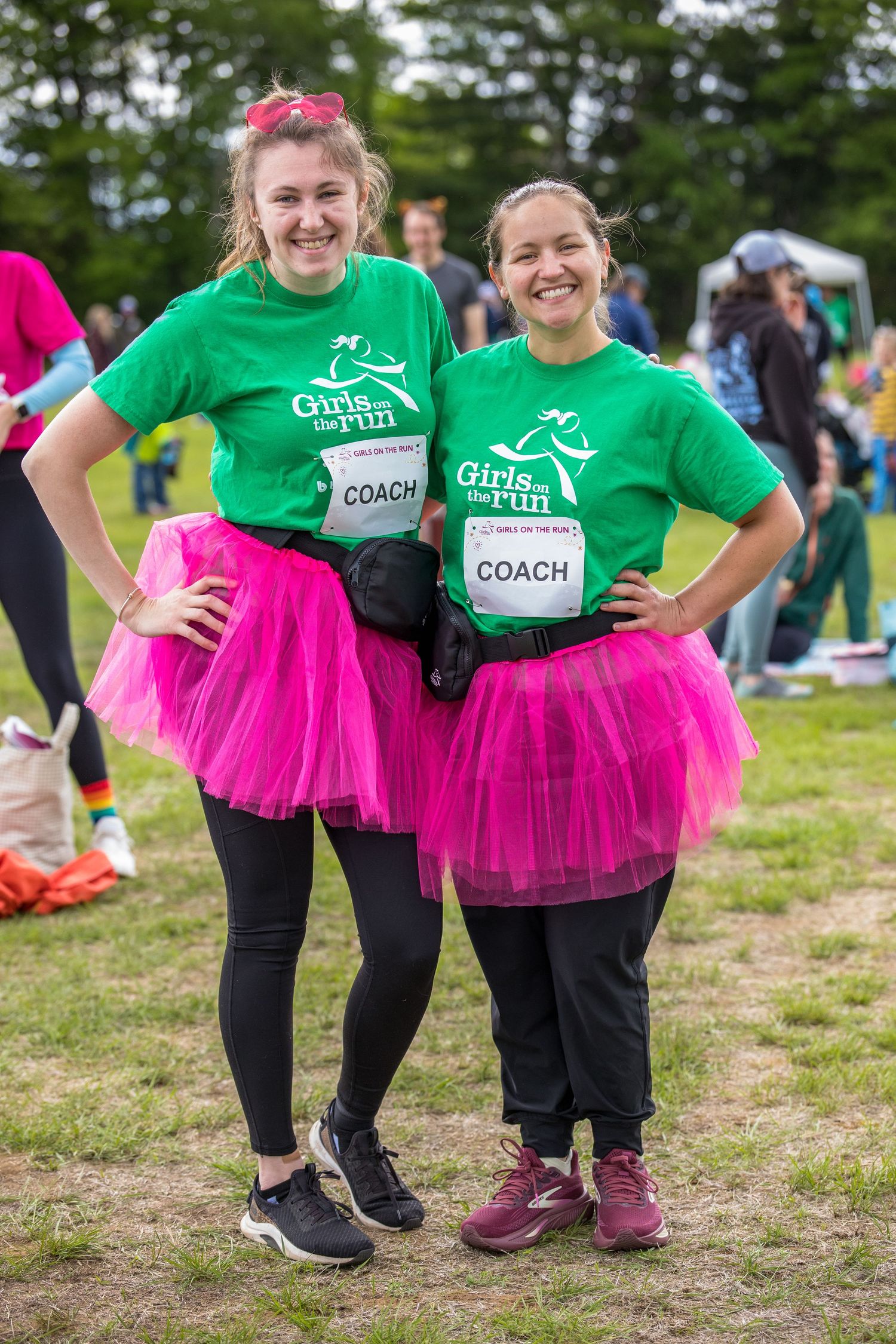 Two women in green Girls on the Run coach shirts and pink tutus pose smiling at an outdoor running event.