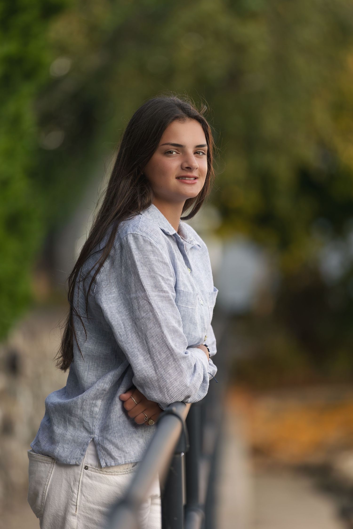 Young woman with long dark hair smiles outdoors, wearing a light blue linen shirt, leaning on a metal railing.