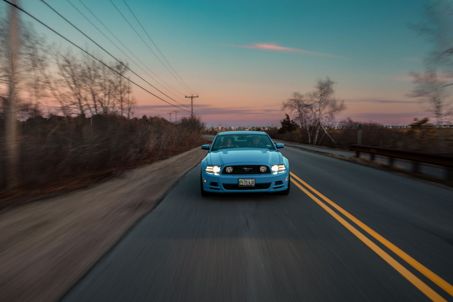 A blue BMW M3 drives on an empty road at sunset with glowing headlights and power lines overhead.