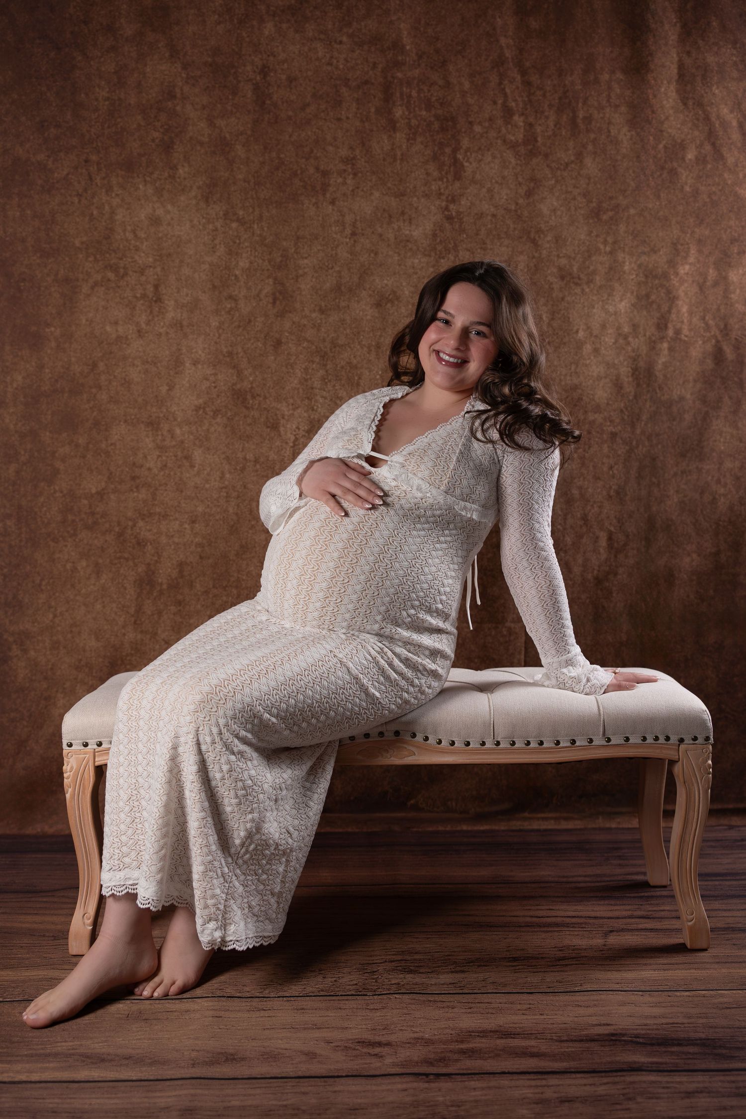 Maternity portrait of an expectant mother in a white lace dress sitting on an elegant bench against a brown backdrop.