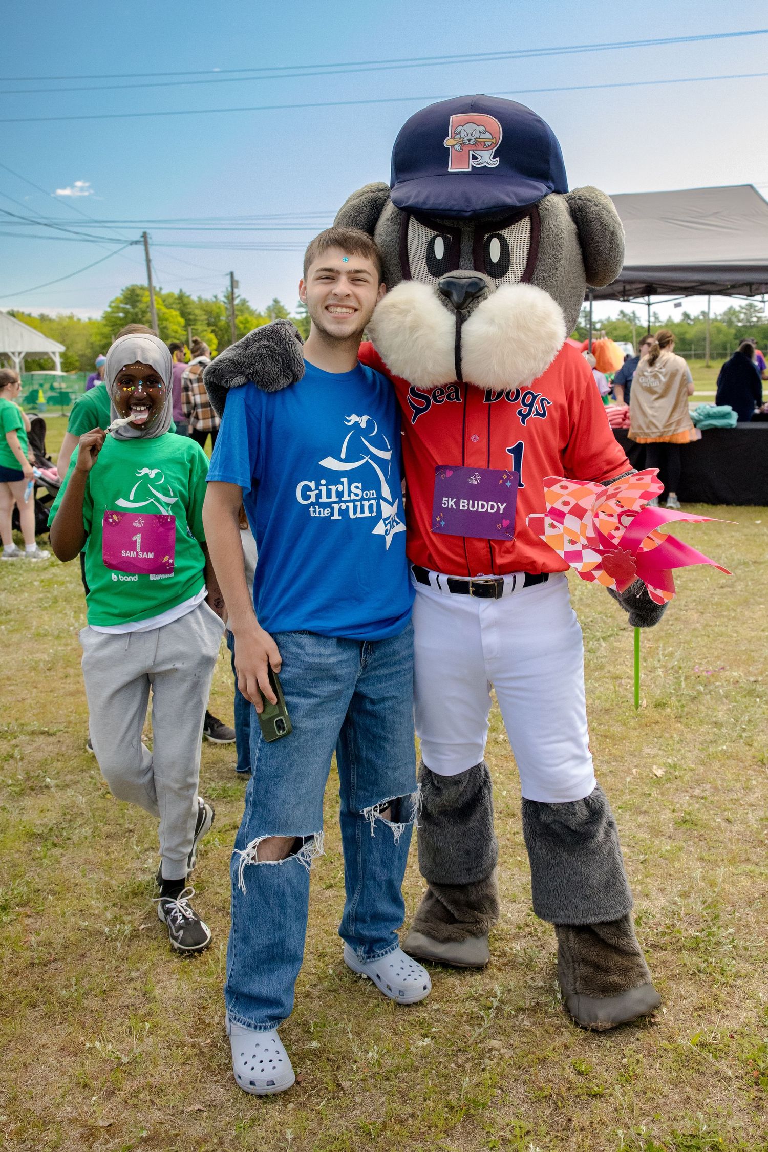 Young man poses with Sea Dogs mascot at Girls on the Run 5K event on sunny day outdoors.