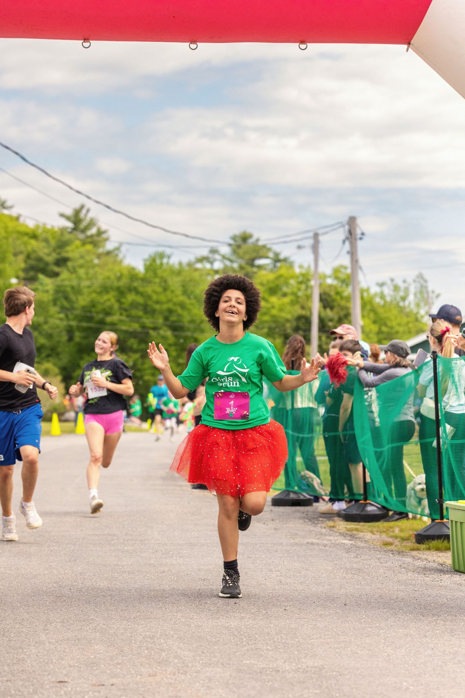 Woman in green shirt and red tutu crosses finish line at outdoor charity fun run on a cloudy day.