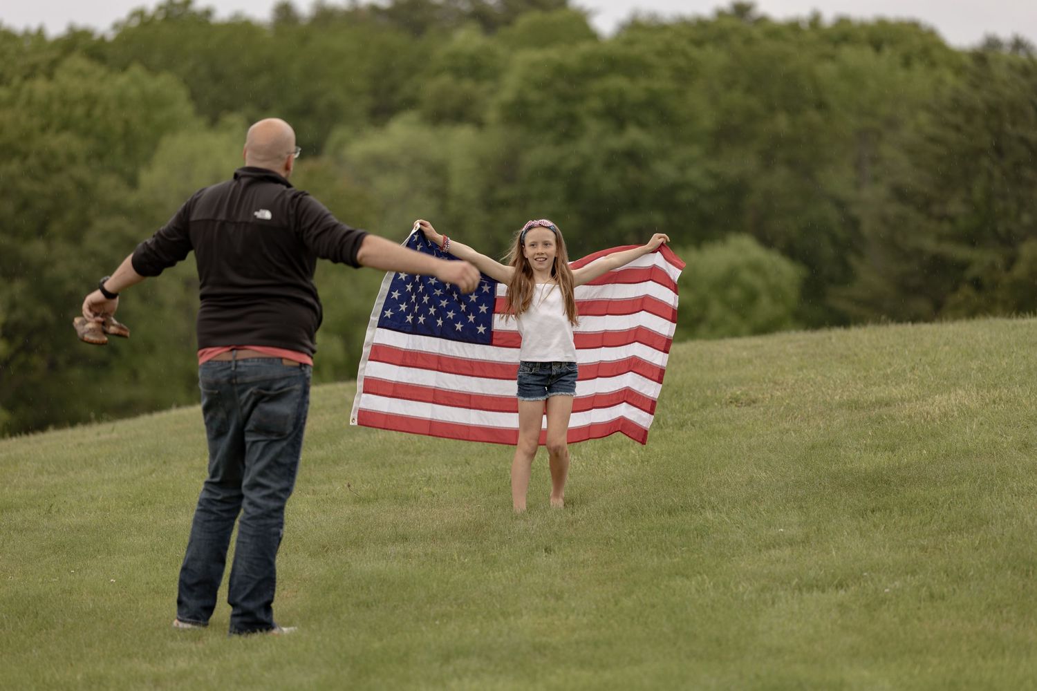 A girl runs toward a man holding an American flag in a green field.