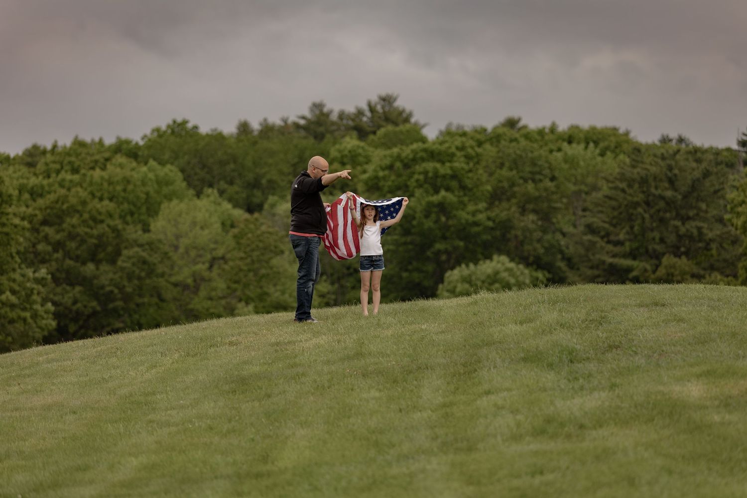 Two people holding an American flag on a green hill with trees in the background on a cloudy day.