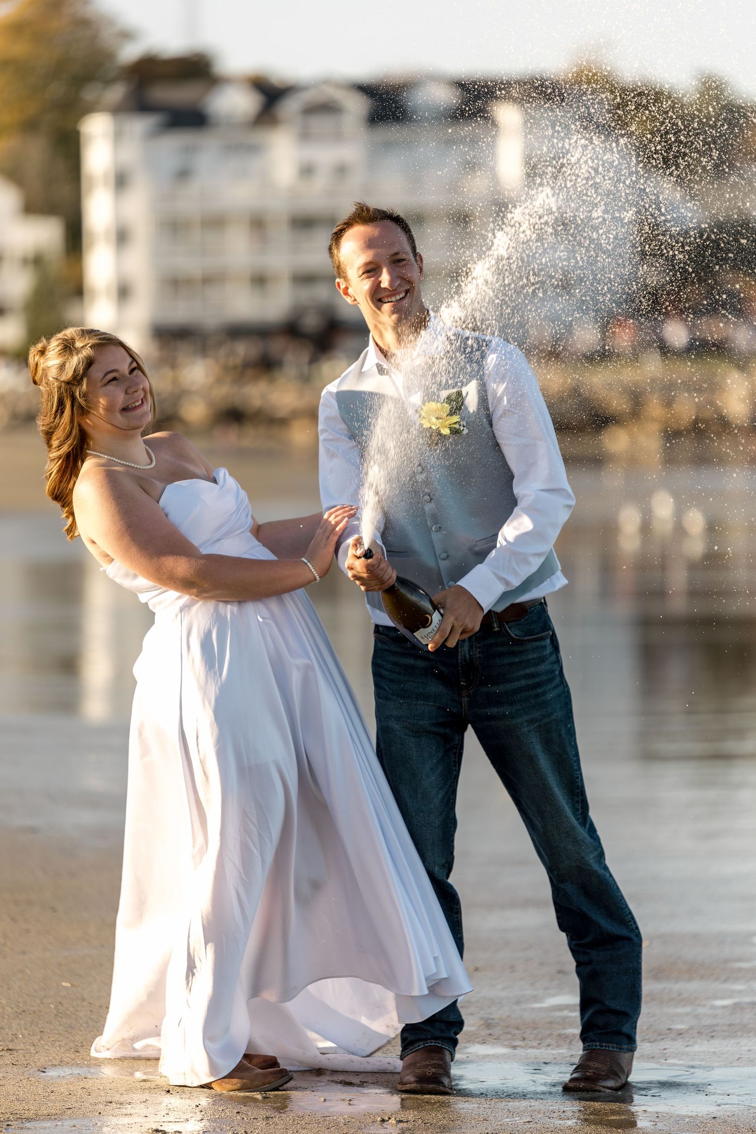 Joyful newlyweds popping champagne on a beach, bride in white gown and groom in jeans, with waterfront building backdrop.