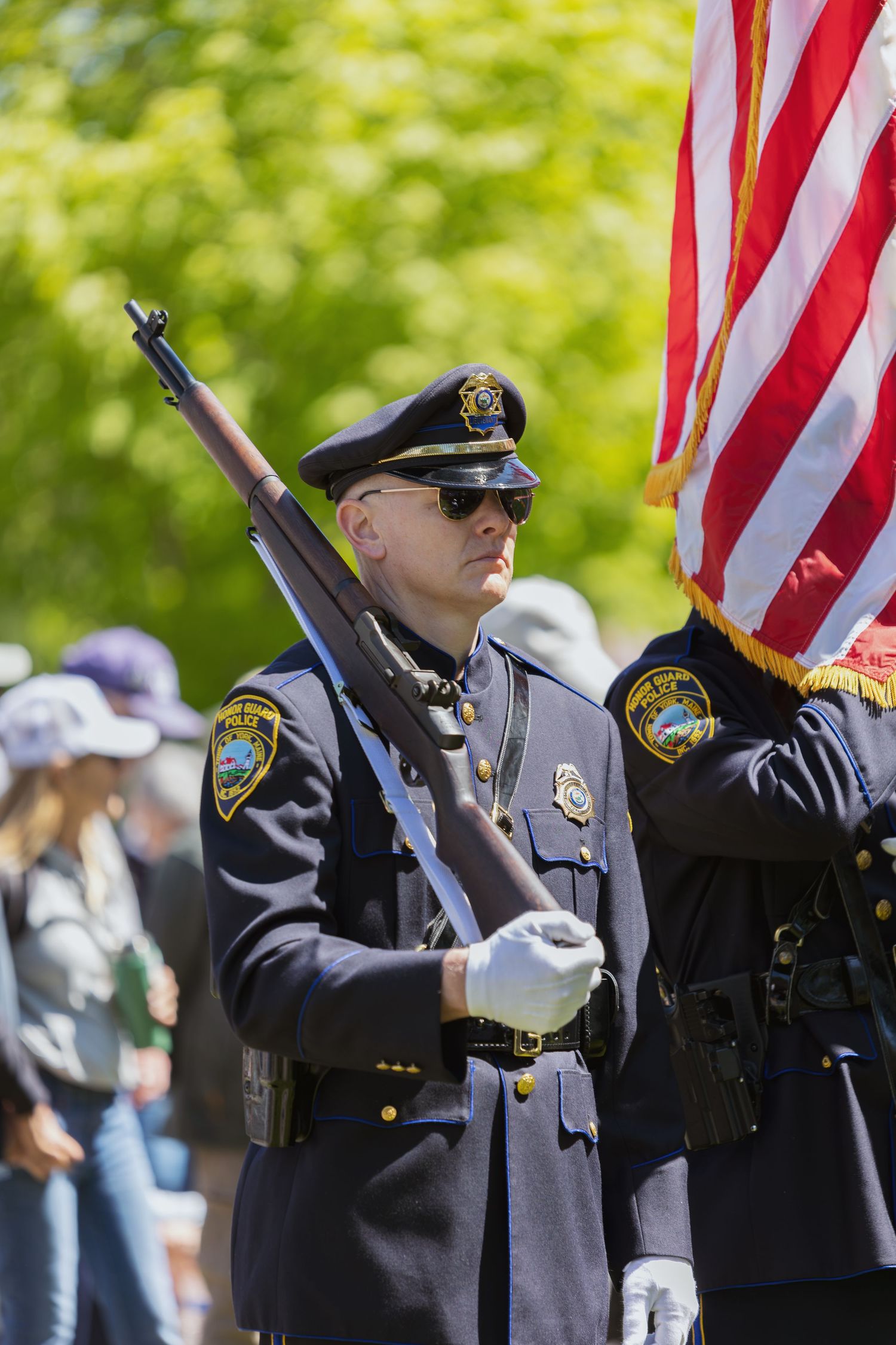Police honor guard in dress uniform holding rifle and American flag at outdoor ceremony.