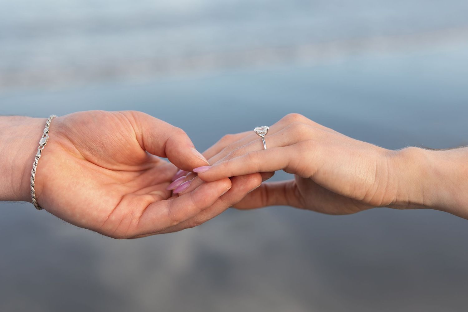 Two hands touching fingers on a beach, one wearing a diamond engagement ring and a silver bracelet.