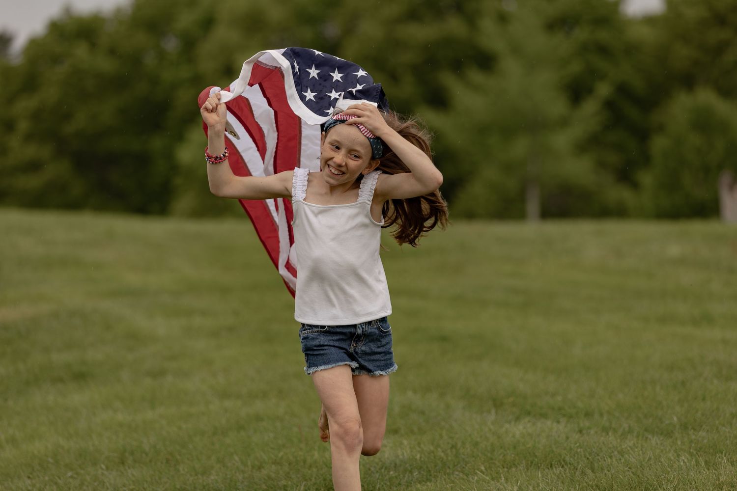 A smiling girl runs through a green park holding an American flag behind her on a sunny day.