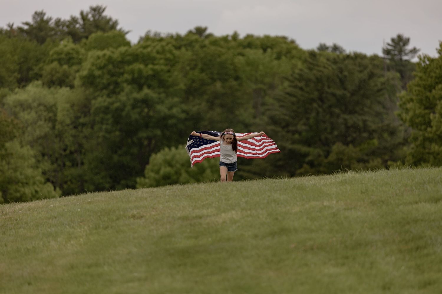 Girl holding an American flag with arms outstretched on a green hill with trees in the background.