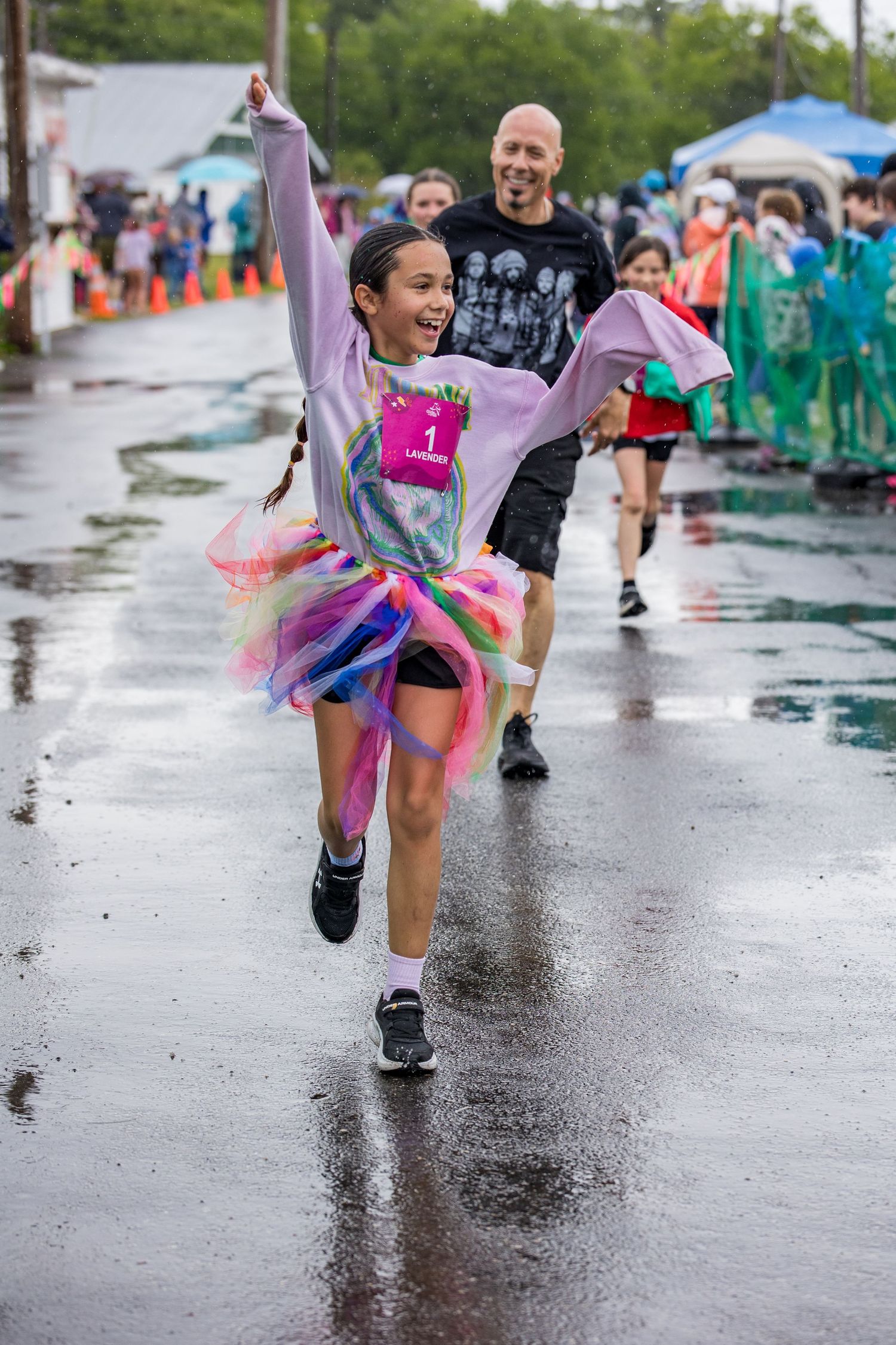 Joyful girl in rainbow tutu crosses finish line at rainy outdoor fun run, arms raised in celebration.