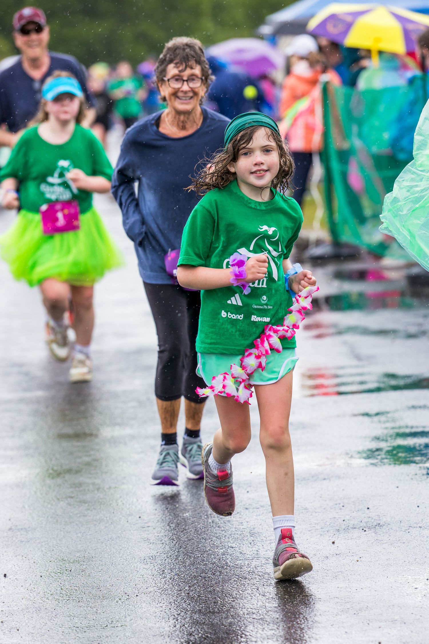 Young girl in green shirt runs joyfully in rainy charity race, with woman and other runners behind her.