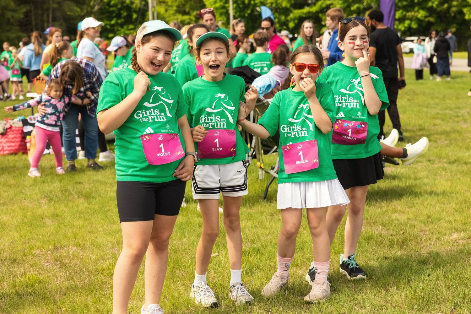 Four girls in green race shirts with pink bibs smile and pose outdoors at a fun run charity event.