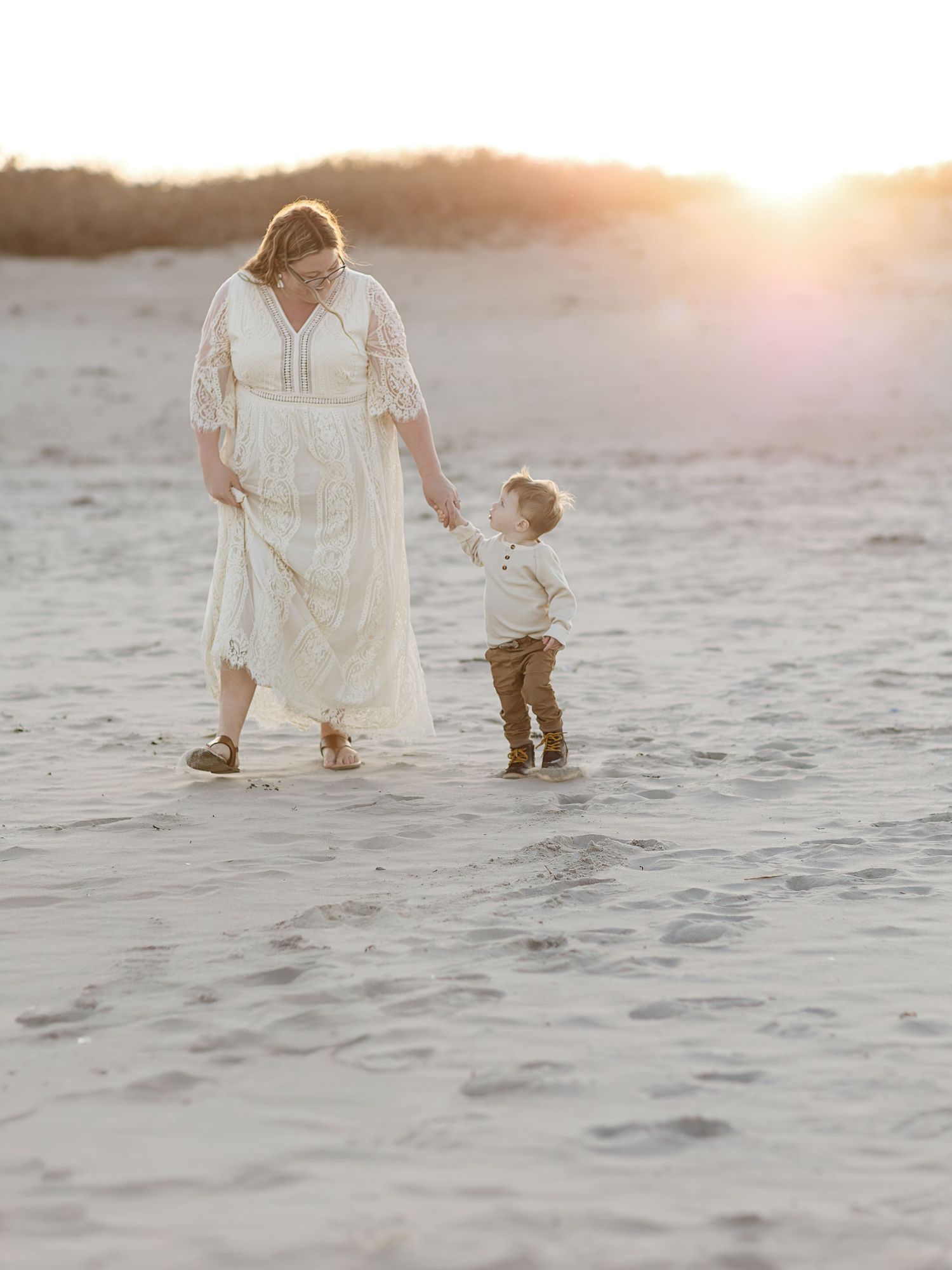 Mother in white lace dress holds toddler son's hand on sandy beach at golden sunset hour.