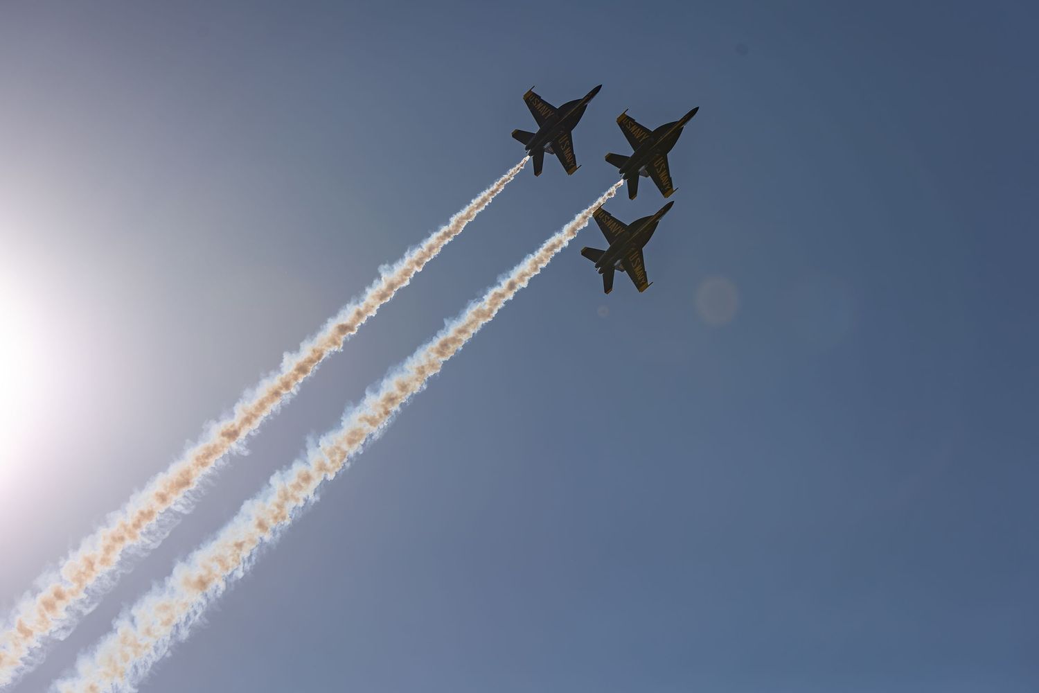 Three military jets fly in tight formation against a clear blue sky, leaving white vapor trails behind them.