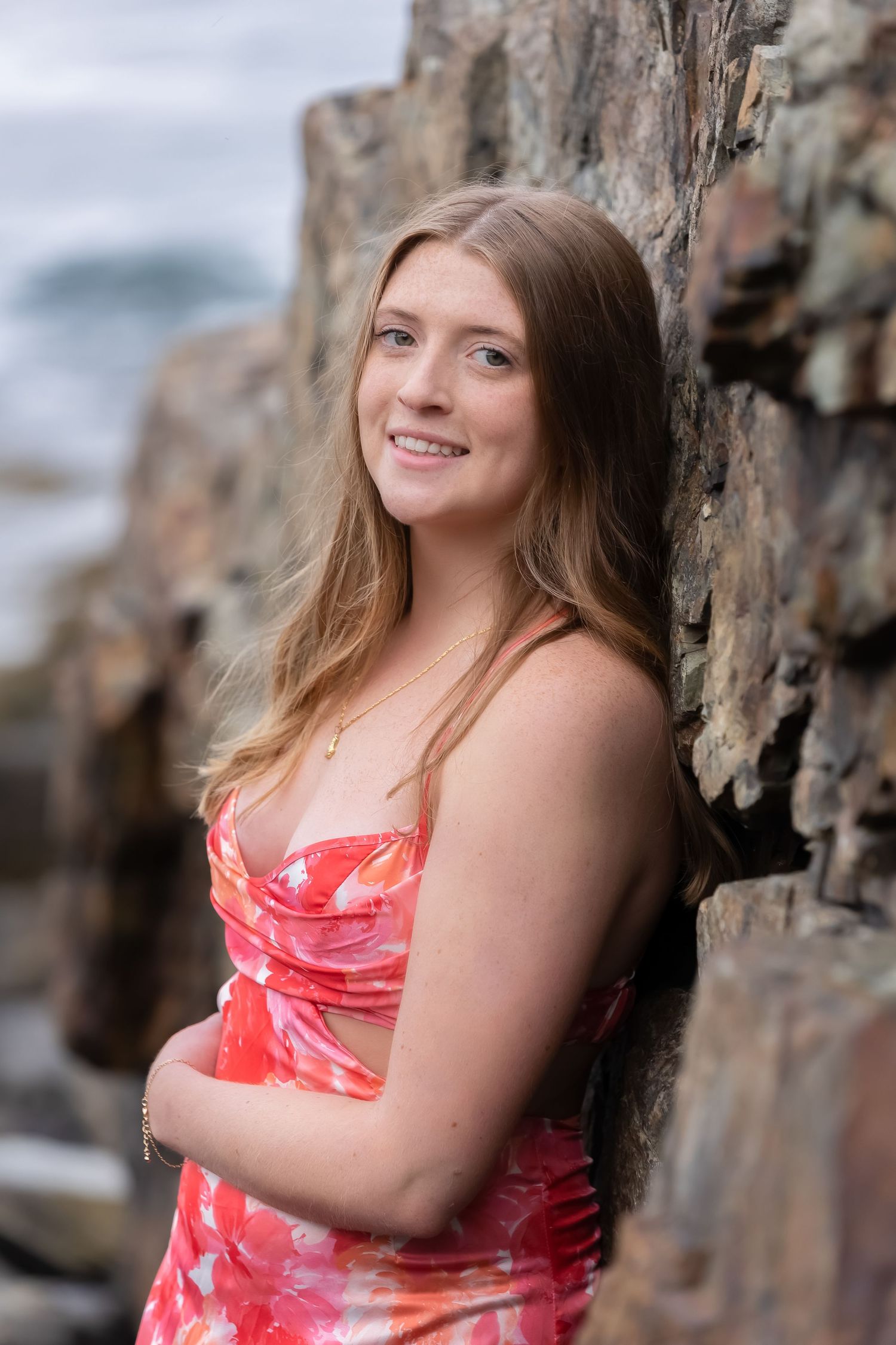 Young woman in floral pink dress leaning against rocky coastal cliffs, smiling outdoors.