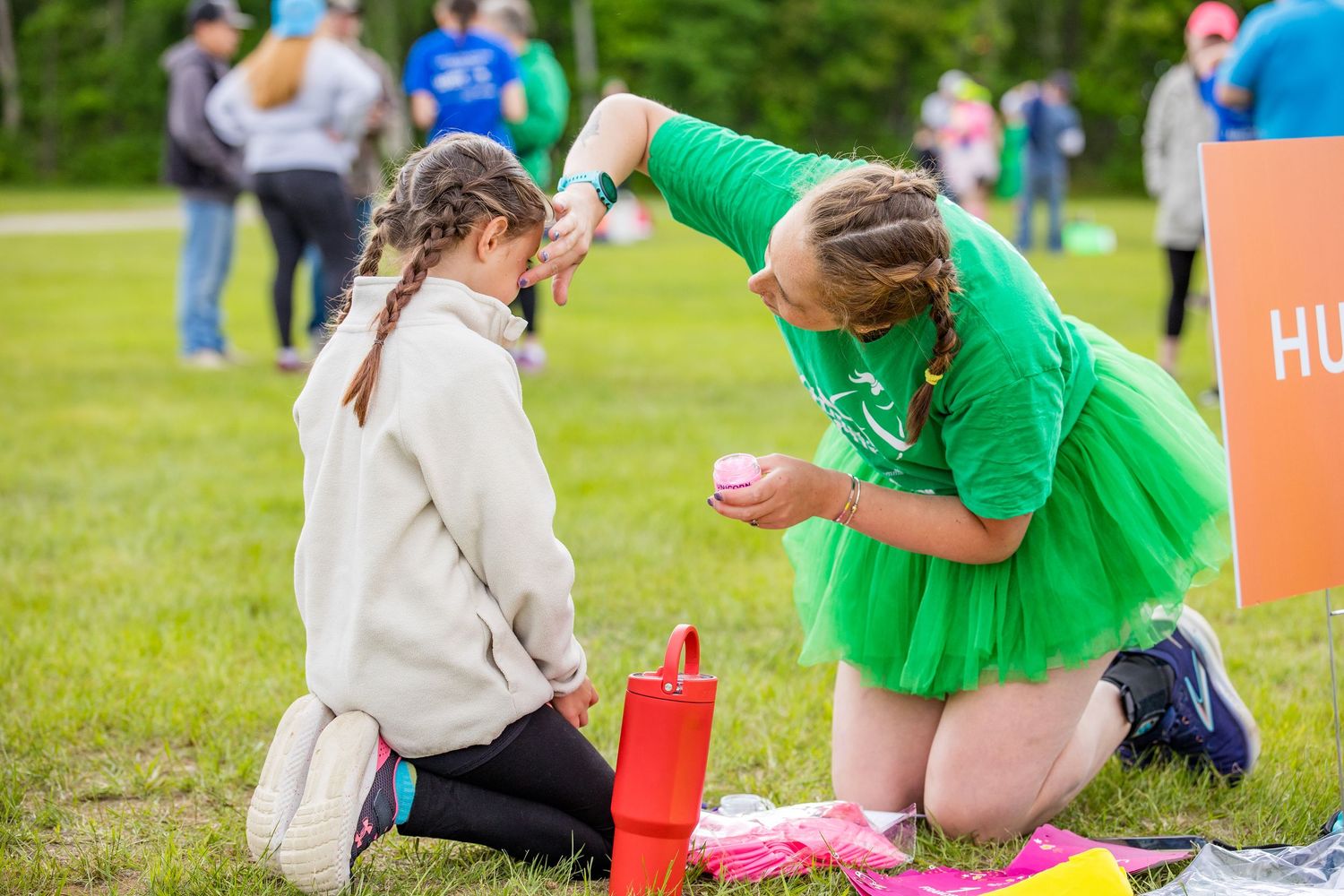 Woman in green tutu applies face paint to a young girl at an outdoor charity fun run event on a grassy field.