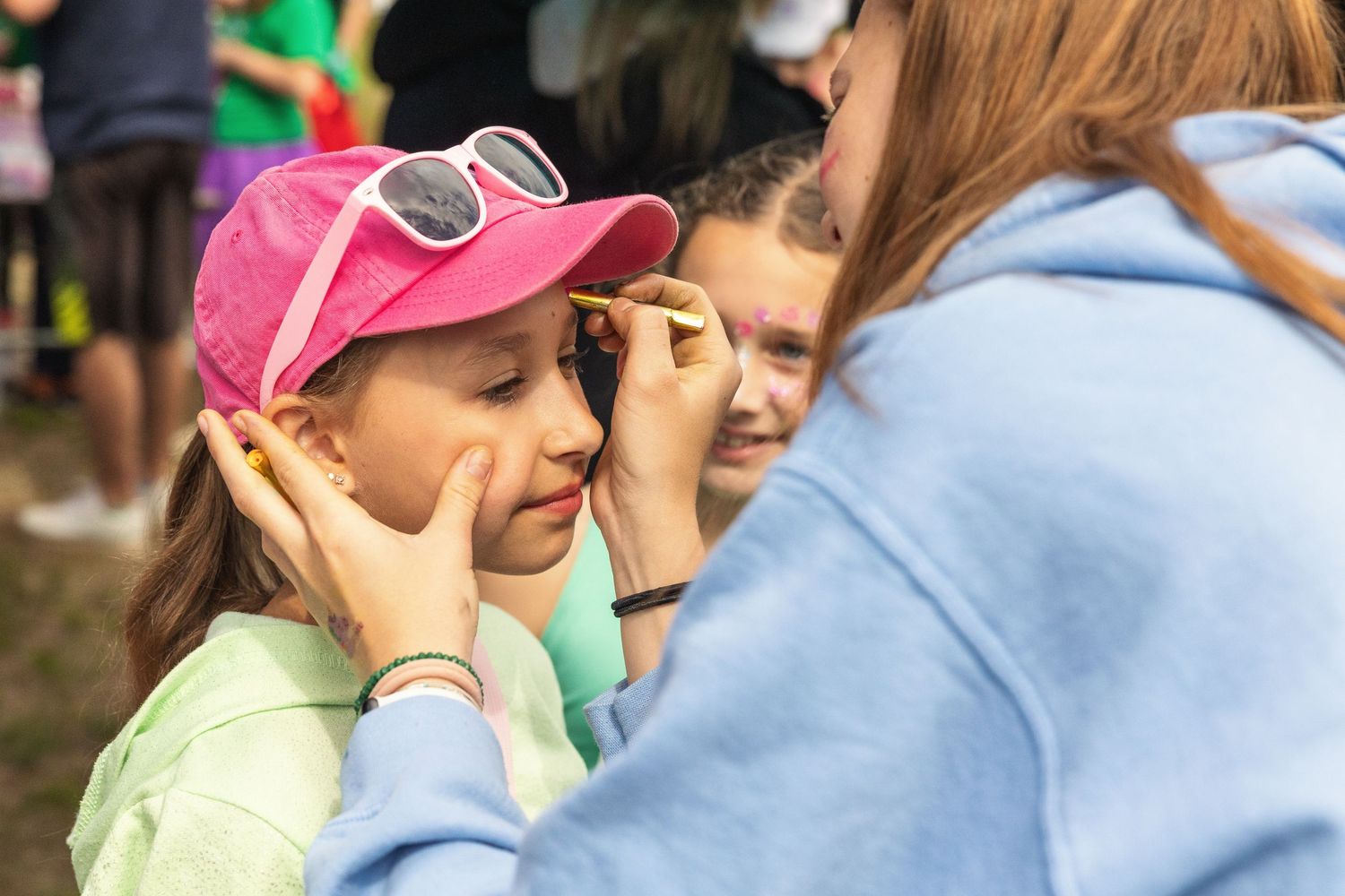 A counselor applies face paint to a young girl in a pink cap at an outdoor camp event.