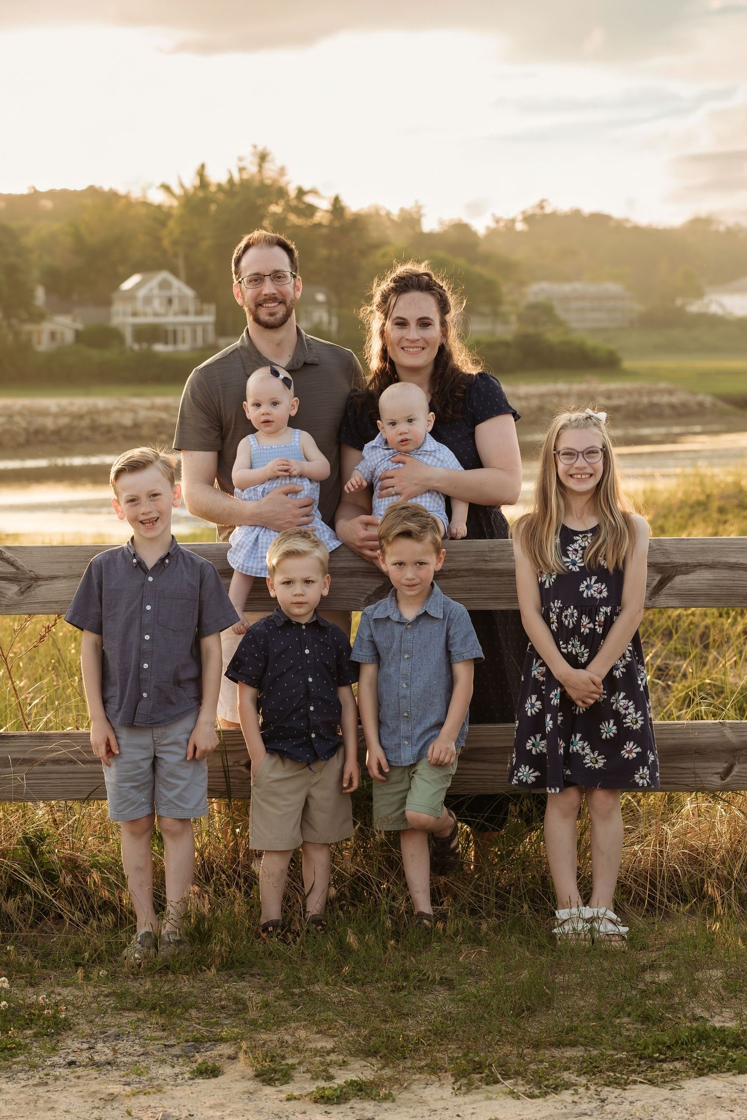 A family poses together on a wooden fence at sunset in coordinated blue and gray outfits during a beach photoshoot.