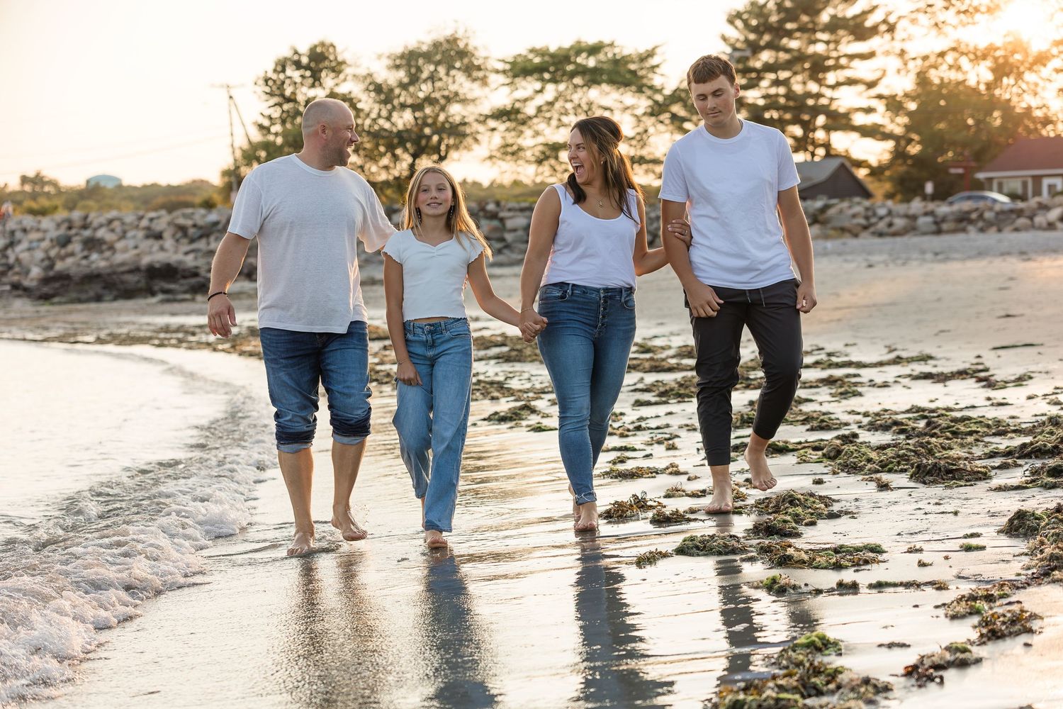 Family of four walking barefoot on a sunny beach at golden hour, holding hands and smiling.