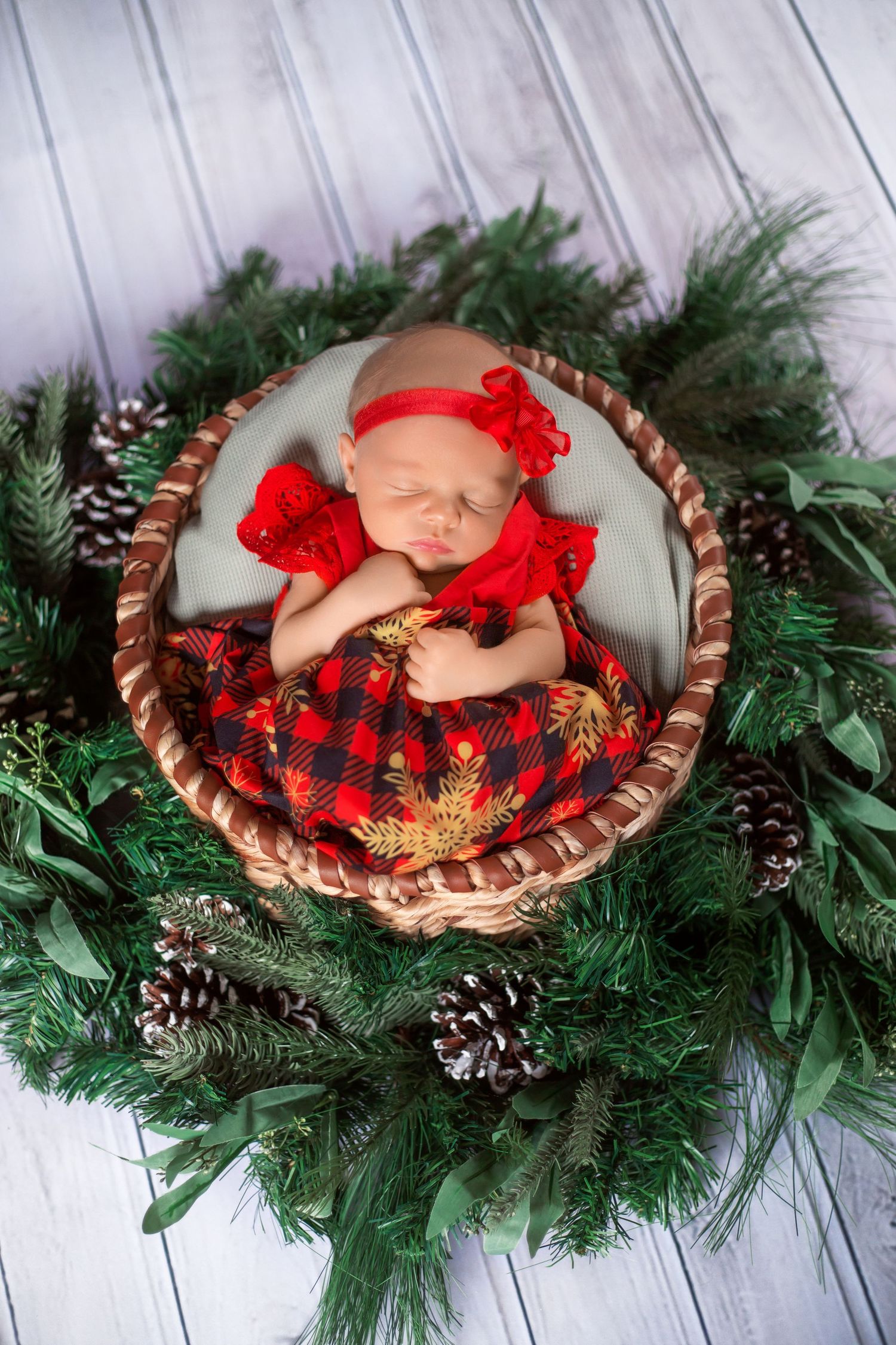 A newborn baby in red plaid holiday attire nestled in a rustic basket surrounded by evergreen branches and pinecones.