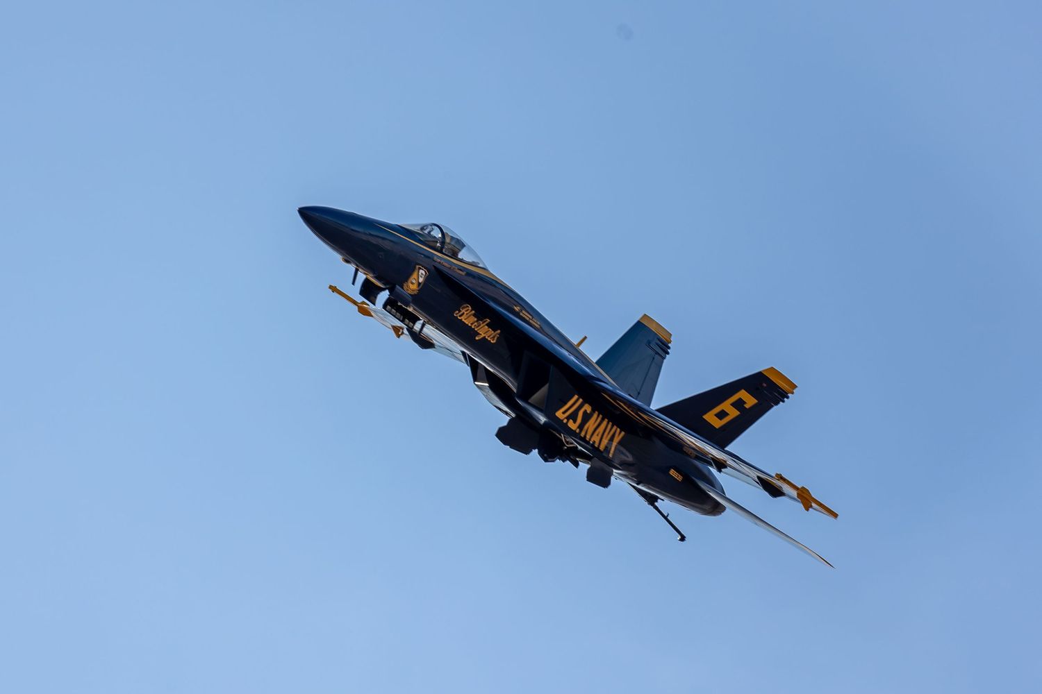 A US Navy Blue Angels F/A-18 Hornet fighter jet performs aerial maneuvers against a bright blue sky.
