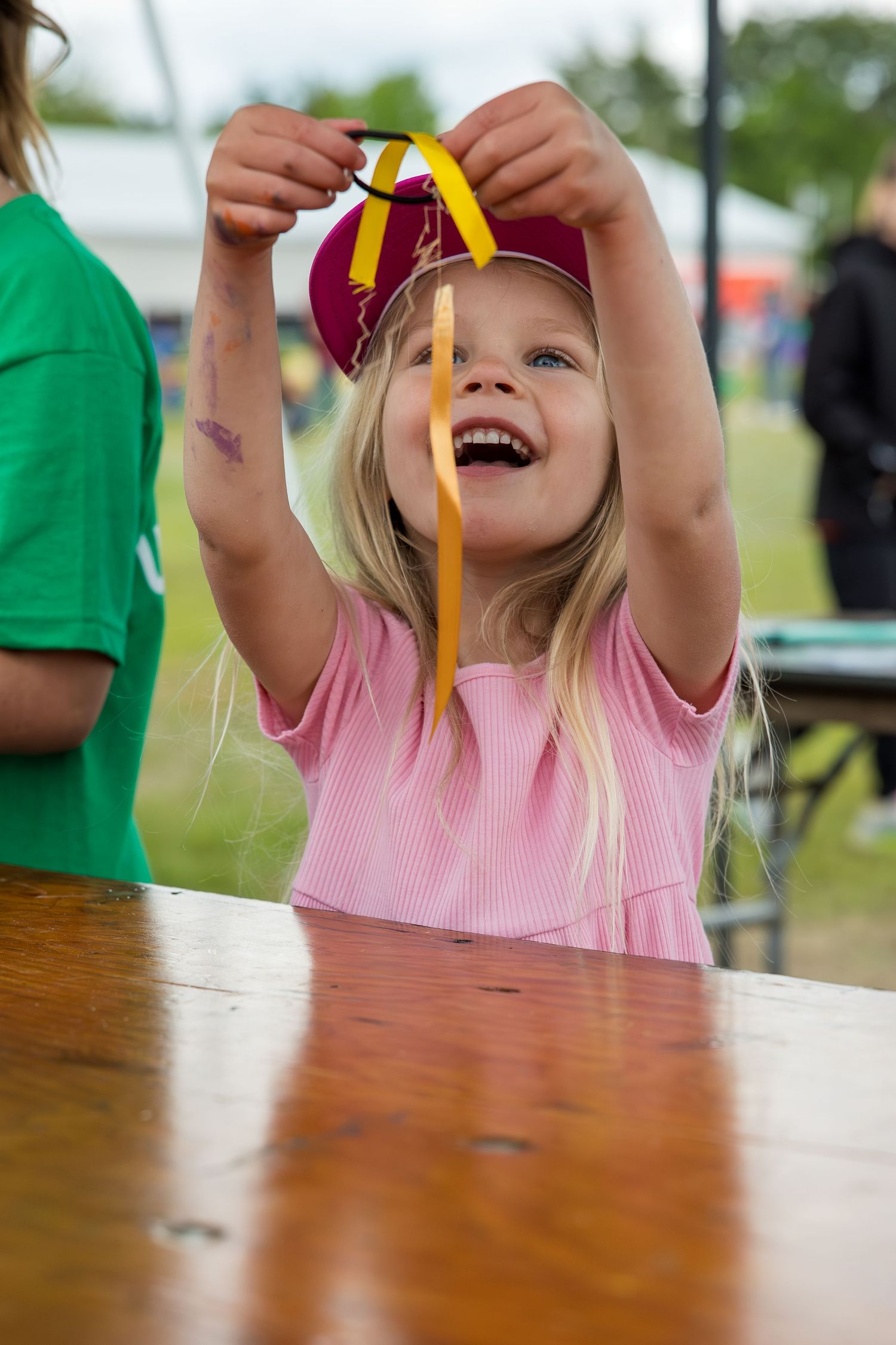 Happy blonde girl in pink shirt holds up a medal ribbon at an outdoor event, laughing with joy.