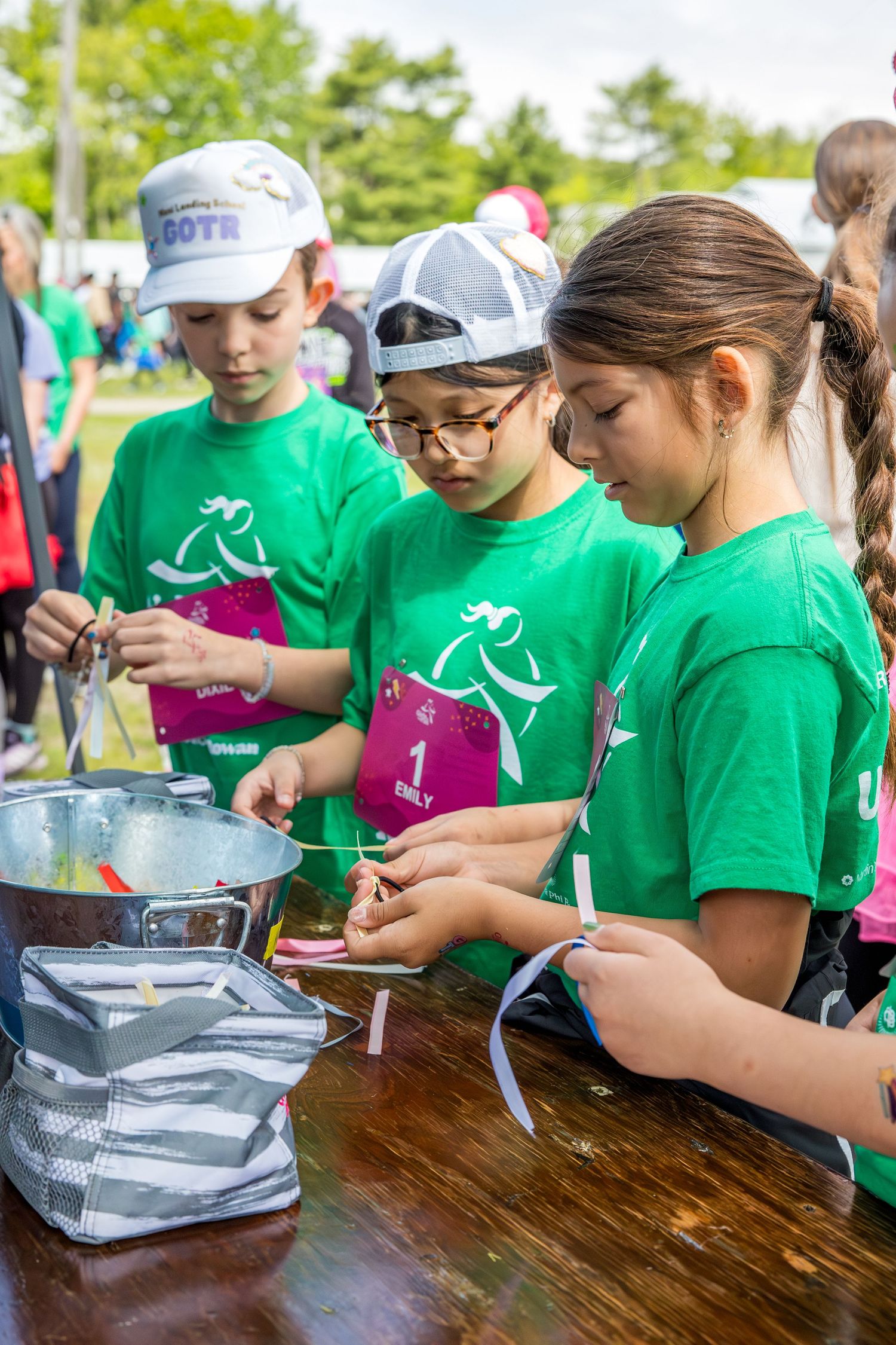 Three children in green t-shirts participate in an outdoor activity at a table during a kids event.
