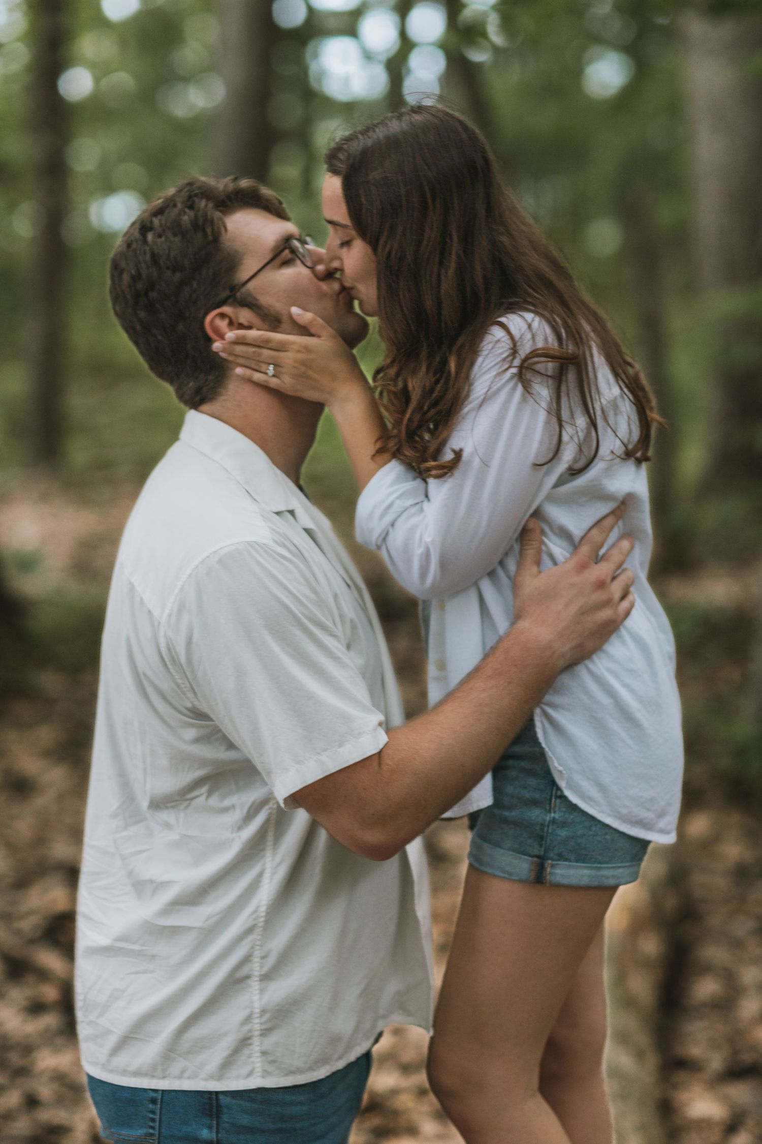 Couple sharing a romantic kiss in a forest setting, both wearing white tops and jeans.