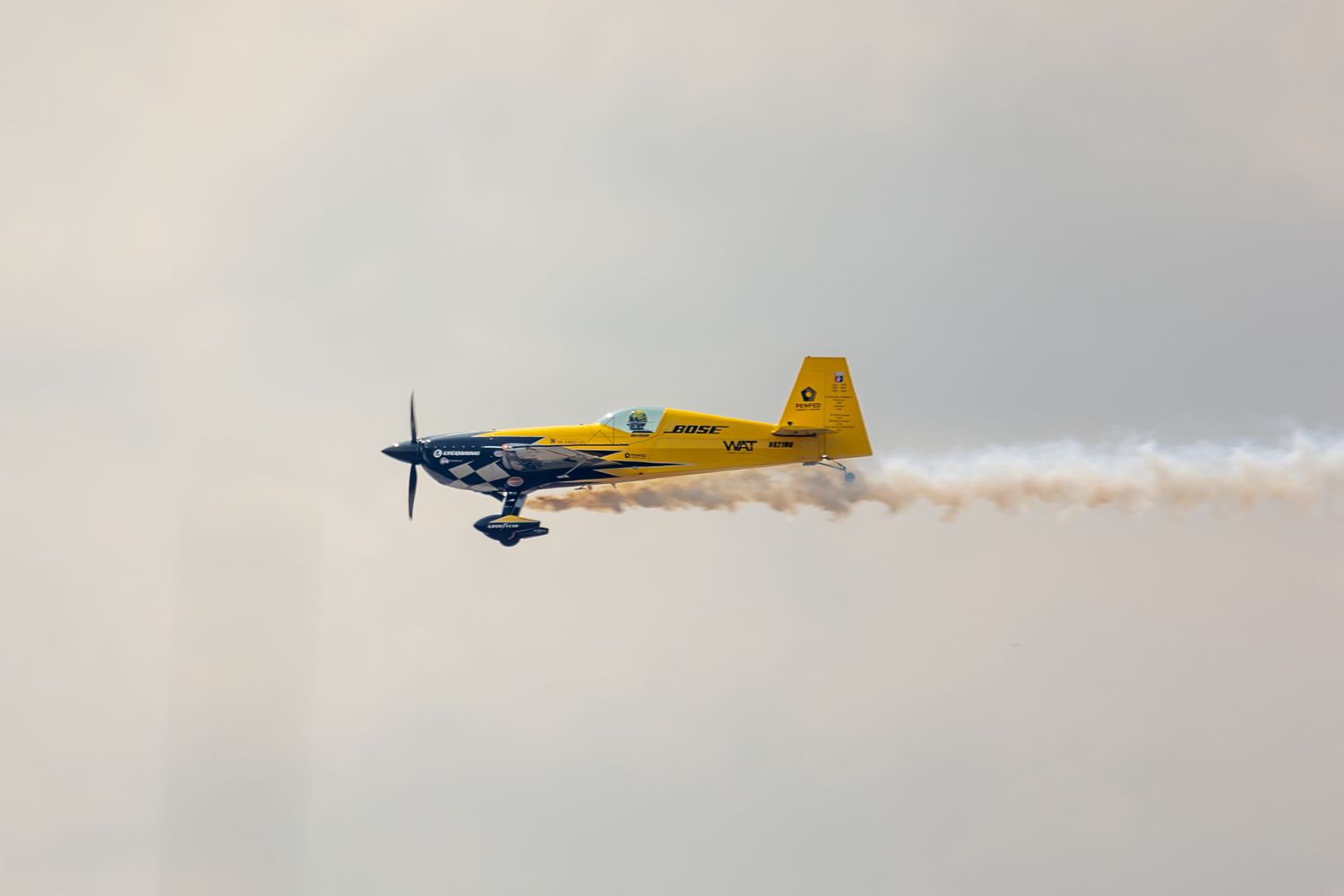 Yellow aerobatic aircraft performs aerial maneuver with white smoke trail against blue sky during airshow.
