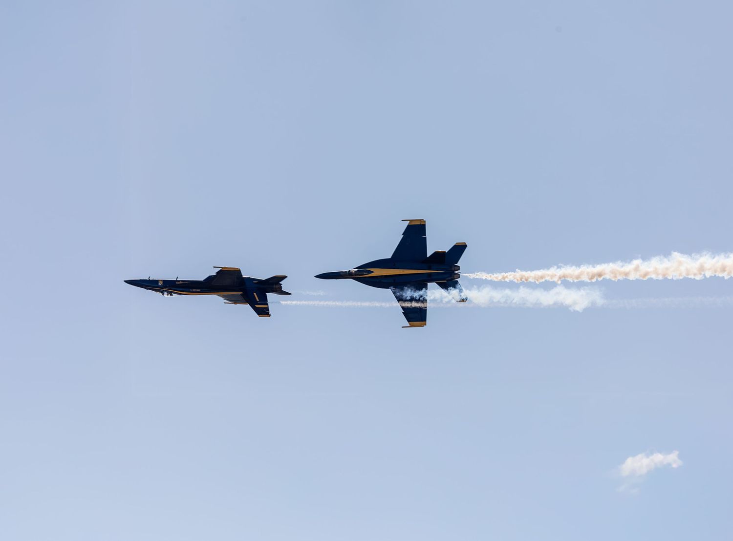 Two Blue Angels F/A-18 Hornet fighter jets perform a high-speed crossing maneuver with white smoke trails in the sky.